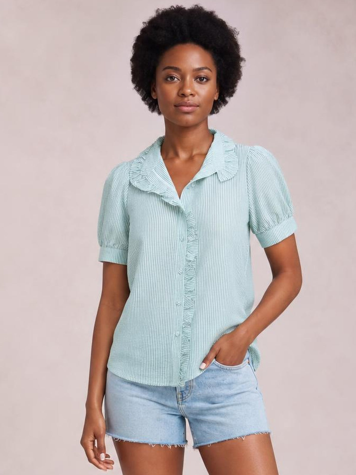 A model wears a light blue striped short sleeve button-down shirt with a ruffled collar and placket, relaxed silhouette, and cuffed sleeves, posing with one hand in her pocket.