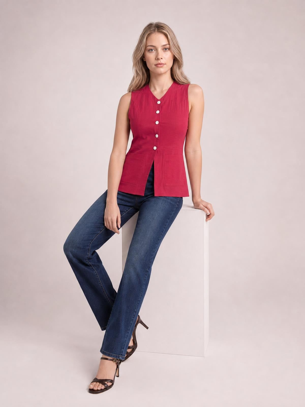 Model seated in a red sleeveless button-down V-neck top with a fitted waist, front pockets, and a straight split hem.