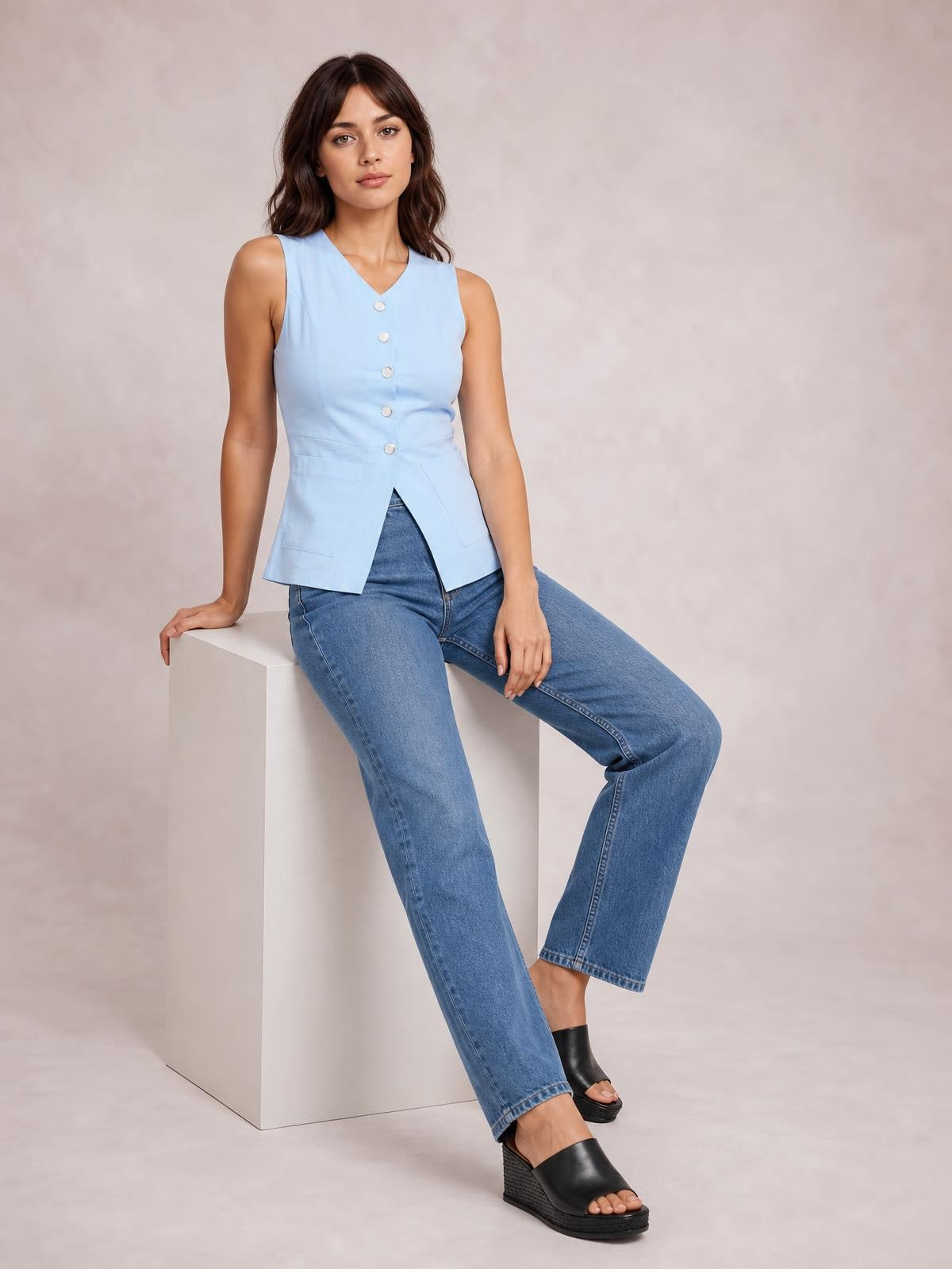 Model seated on a cube wears a light blue sleeveless V-neck top with button-down front, fitted waist, and pointed hem with visible pockets.