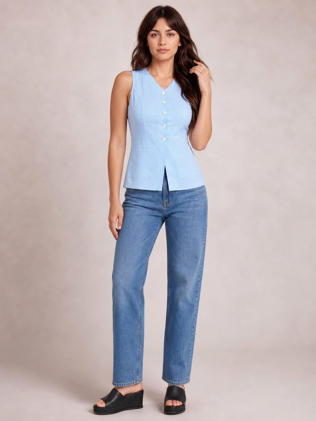 Model wearing a light blue sleeveless button-down V-neck top with a fitted waist, front pockets, and straight hem, posed with one hand touching her hair.