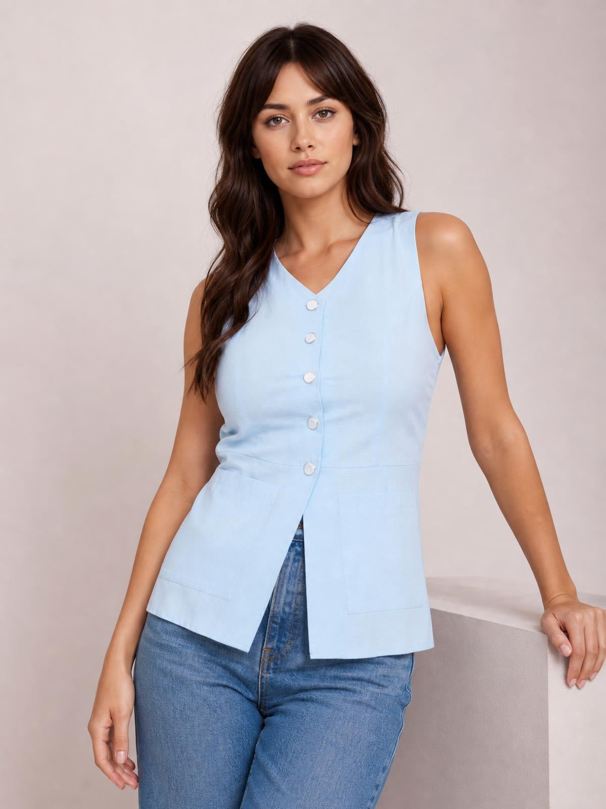 Model wearing a light blue button-down V-neck sleeveless top with a tailored silhouette, front buttons, and two large patch pockets, standing in jeans.