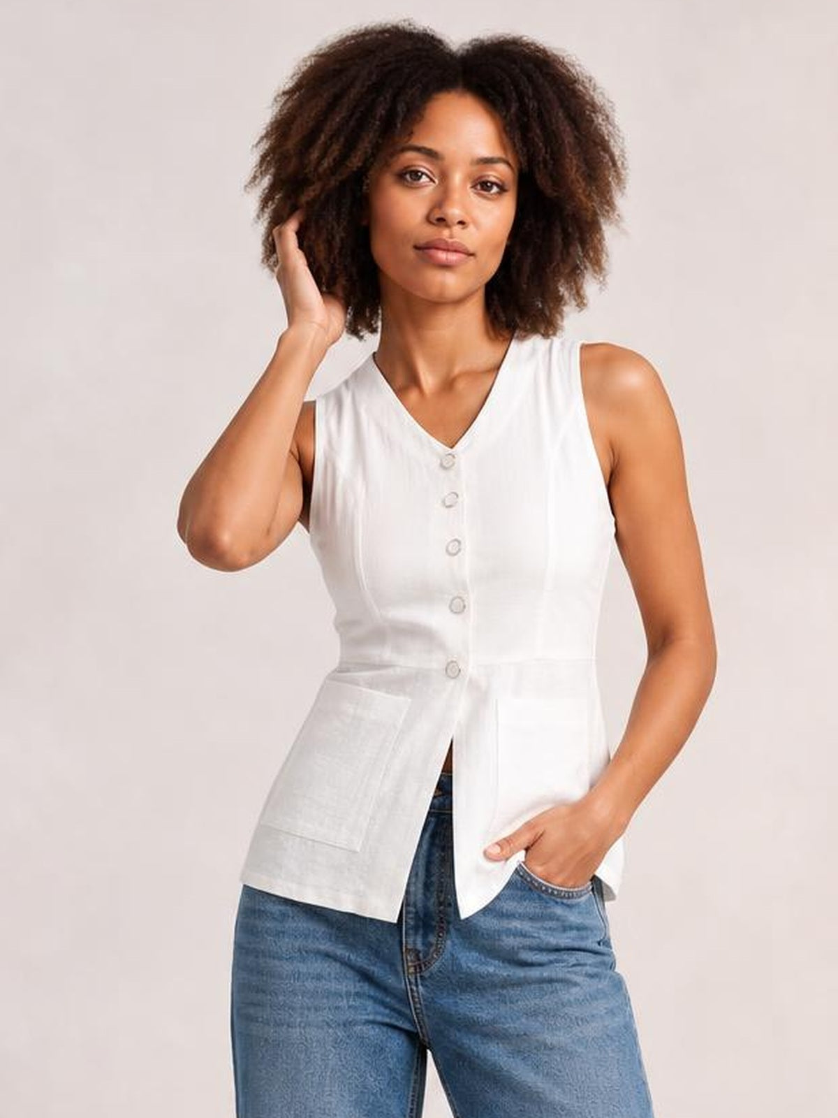 Model wearing a white sleeveless button-down V-neck top with a fitted waist, front buttons, and large patch pockets, posing with one hand in her jeans pocket.