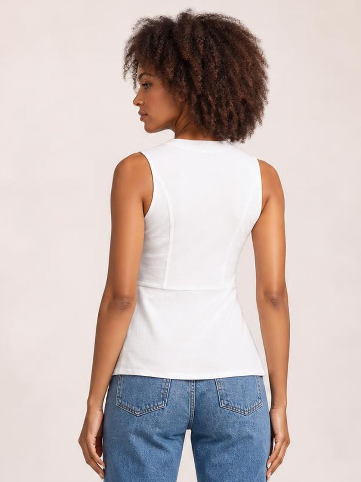 A model looks over her shoulder wearing a white sleeveless top with a fitted silhouette, clean back construction, and a visible waist seam.