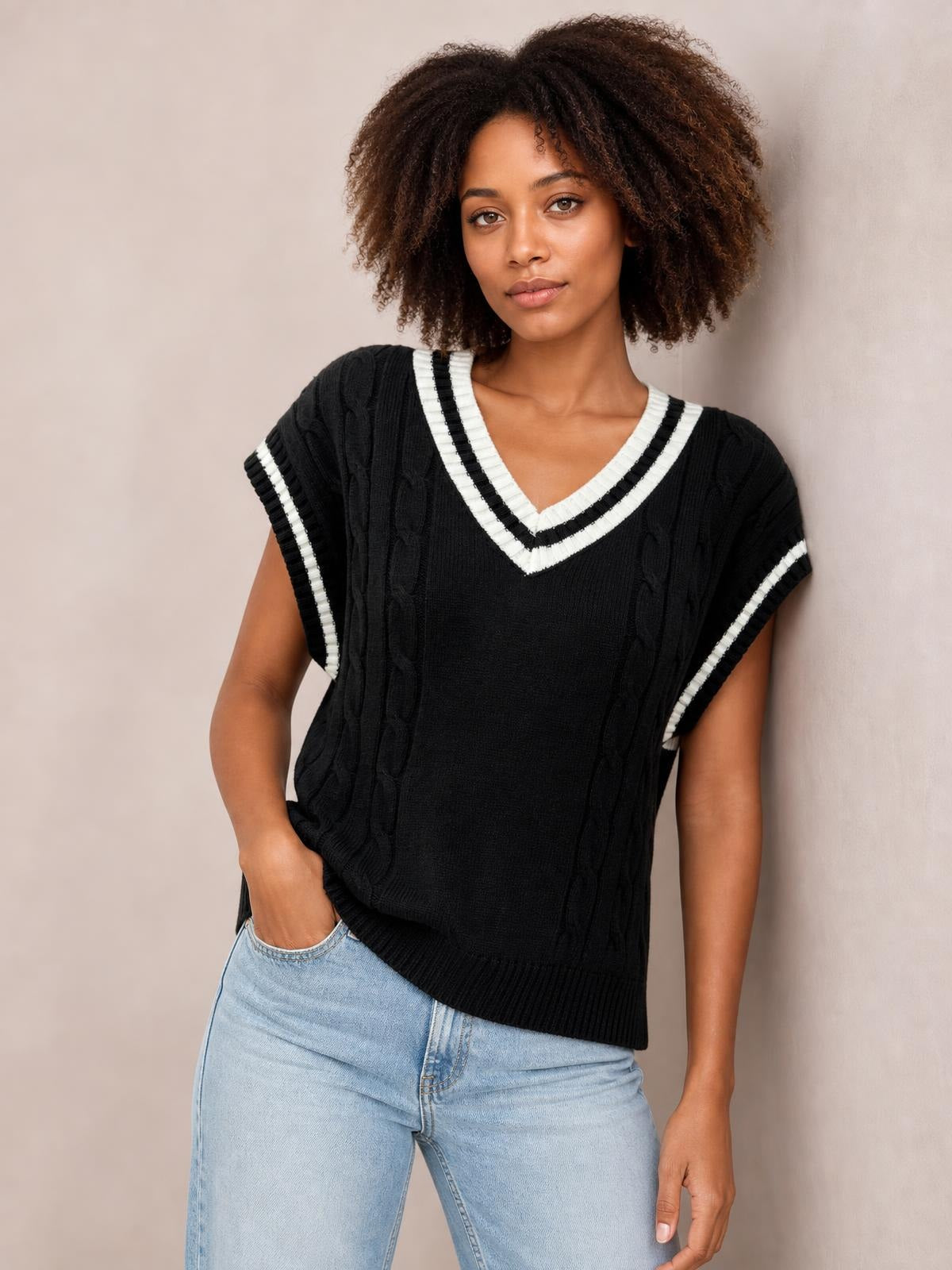 Model poses with one hand in her jeans pocket wearing a black cable-knit V-neck sweater vest with striped trim, ribbed edges, and a relaxed silhouette.