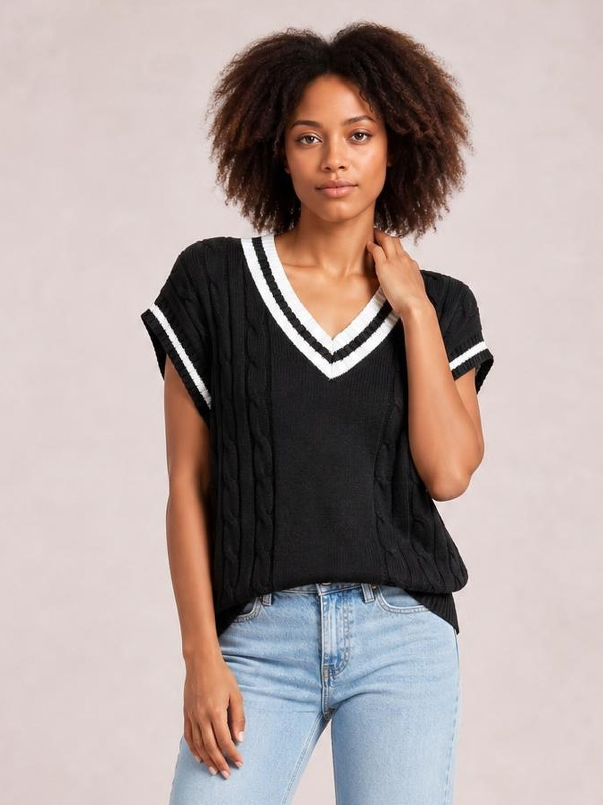 A model wears a black cable-knit V-neck sweater vest with white striped trim, a relaxed silhouette, short sleeves, and one hand resting near the neckline.