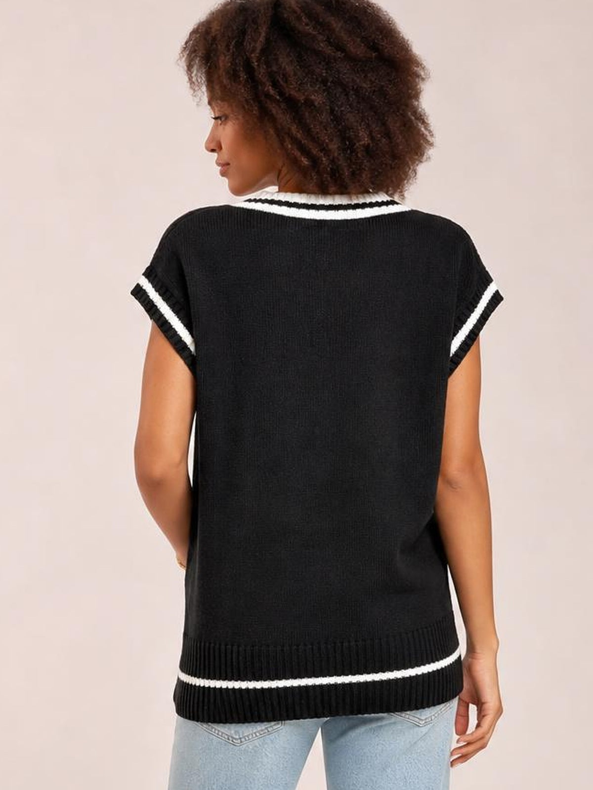 A model shown from the back wears a black knit sweater vest with a relaxed silhouette, ribbed hem, and contrasting white stripes at the neckline, sleeves, and waist.
