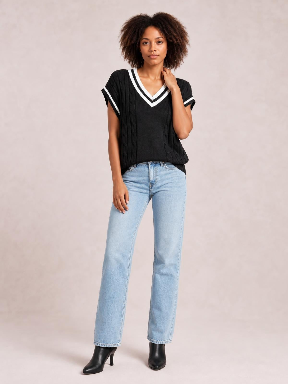 Model poses in a black cable-knit V-neck sweater vest with white trim, short sleeves, a relaxed silhouette, and ribbed edges.
