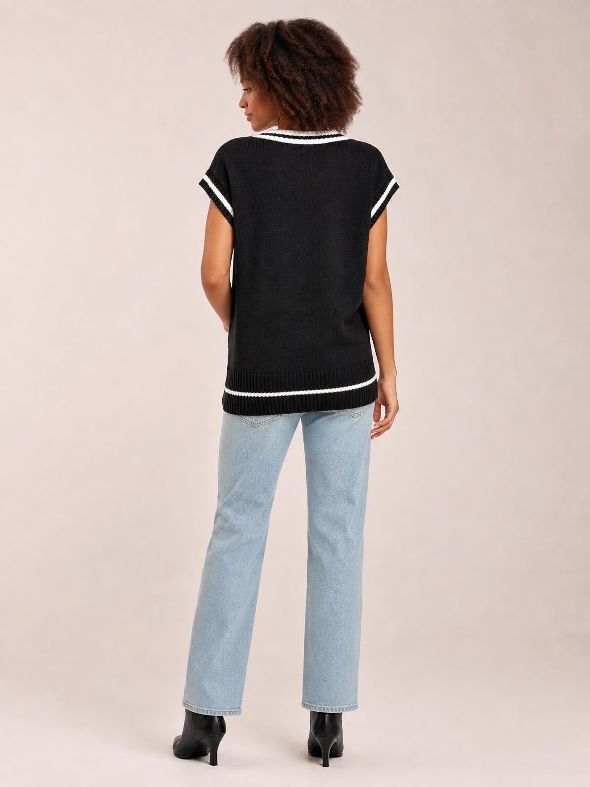 A model stands facing away in a black sweater vest with a relaxed longline silhouette, ribbed edges, and contrasting white stripes at the armholes and hem.