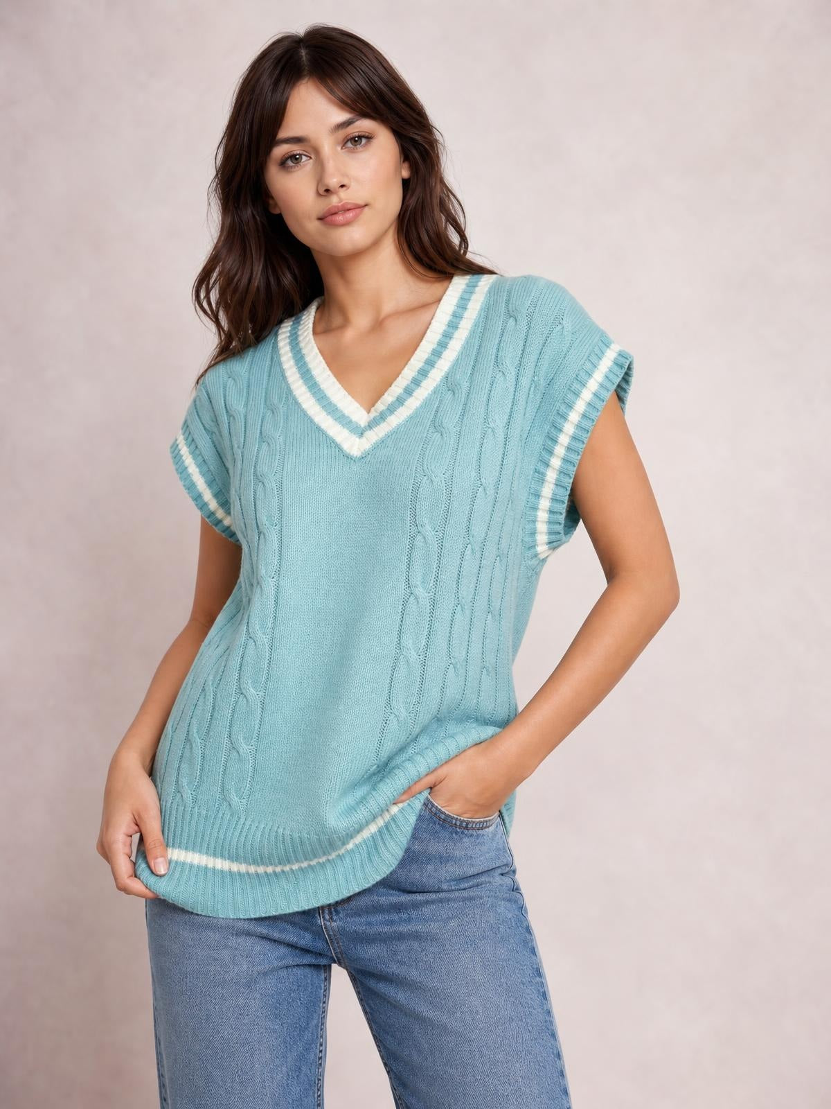 Model poses in a light blue cable-knit V-neck sweater vest with white striped trim, relaxed sleeveless silhouette, and ribbed hem over jeans.