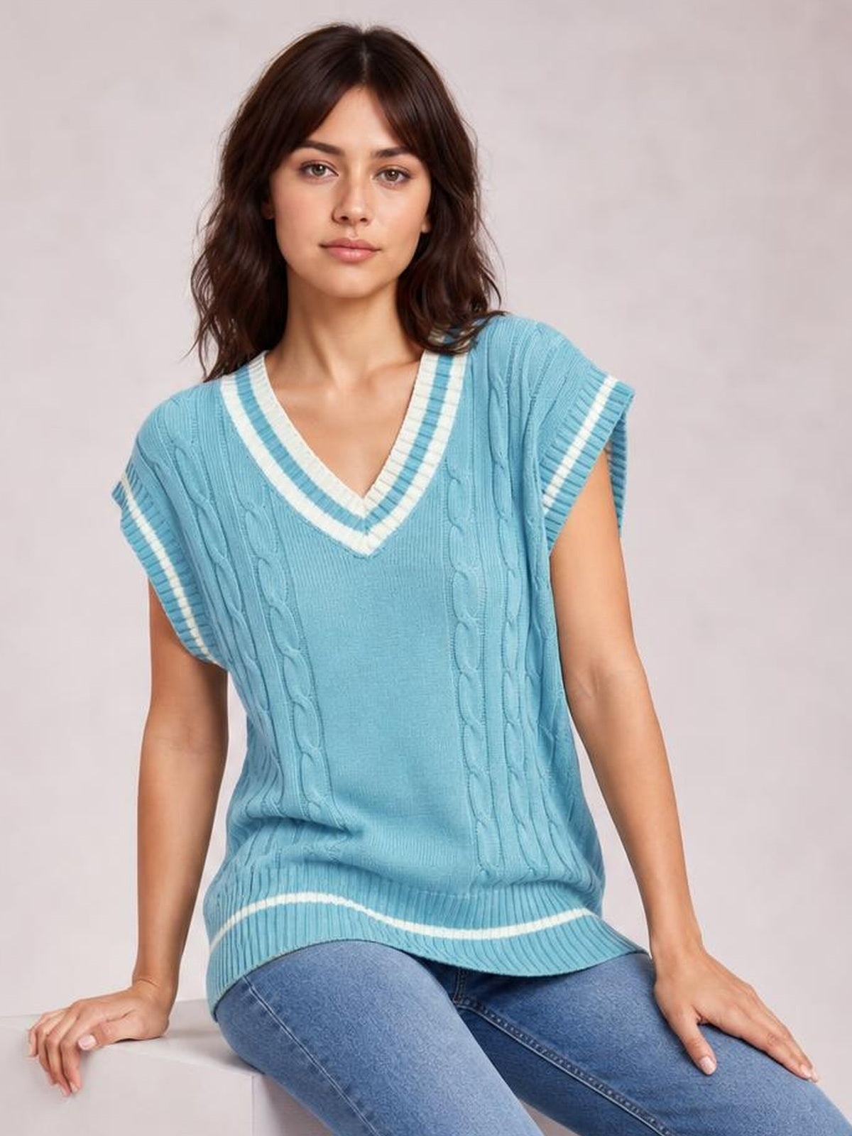 A seated model wears a light blue cable-knit V-neck sweater vest with white striped trim, cap sleeves, a relaxed silhouette, and ribbed hem.