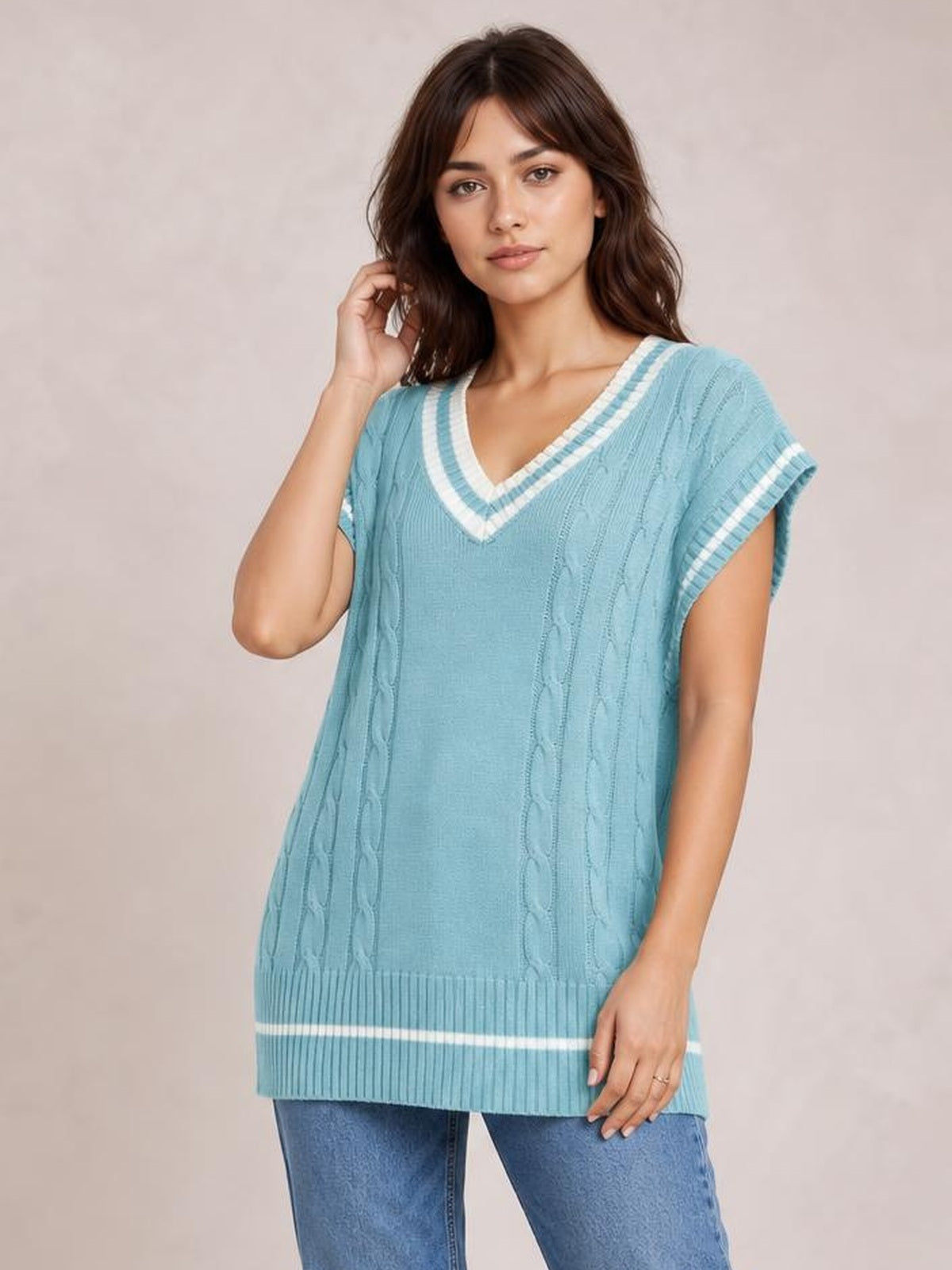 Model poses in a light blue cable-knit V-neck sweater vest with striped ribbed trim, relaxed longline silhouette, and cap sleeves.