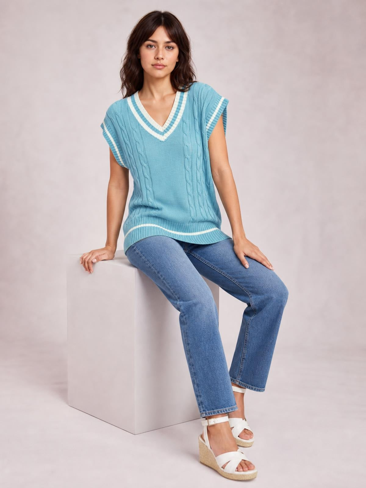 A model sits wearing a light blue cable-knit V-neck sweater vest with white striped trim, relaxed sleeveless silhouette, and ribbed hem and armholes.