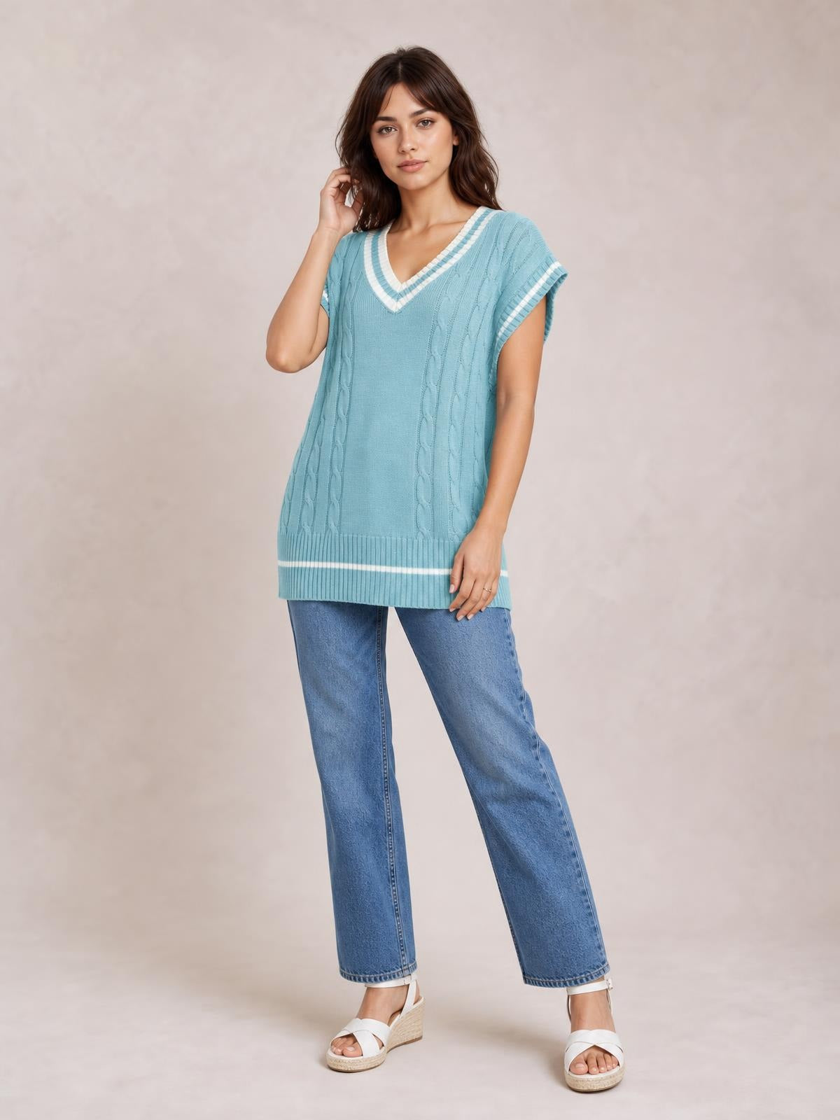 A model poses in a relaxed stance wearing a light blue cable-knit V-neck sweater vest with white trim, ribbed edges, and a long relaxed silhouette.