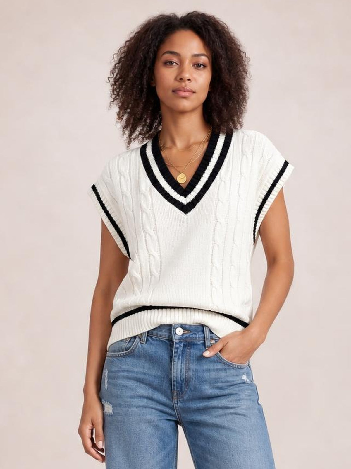 Model wearing a white cable-knit V-neck sweater vest with black striped ribbed trim, sleeveless silhouette, and a relaxed hem over jeans.