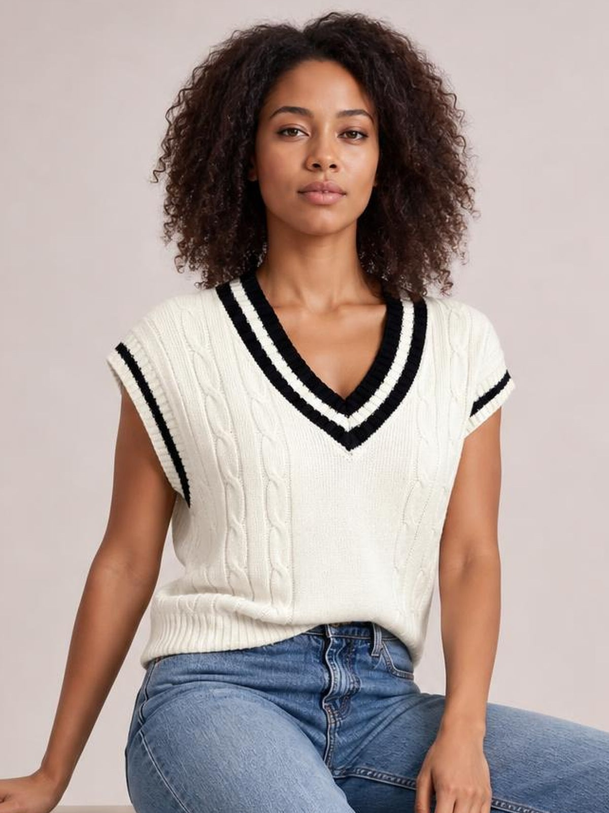 A model sits wearing a cream cable-knit V-neck sweater vest with black striped trim, sleeveless construction, and a relaxed straight silhouette.