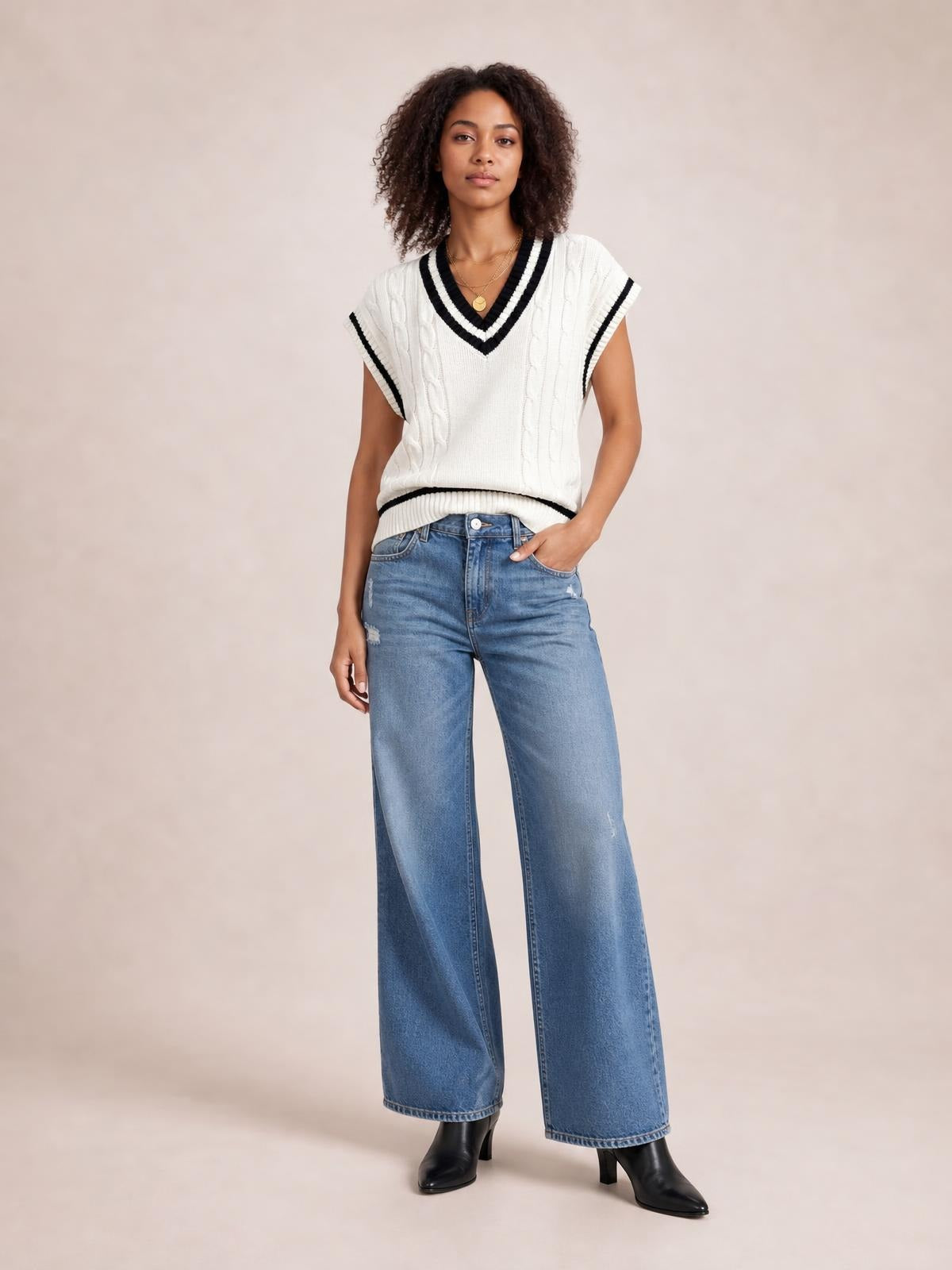 Model poses in a white cable-knit V-neck sweater vest with black trim, ribbed edges, short sleeves, and a relaxed silhouette.