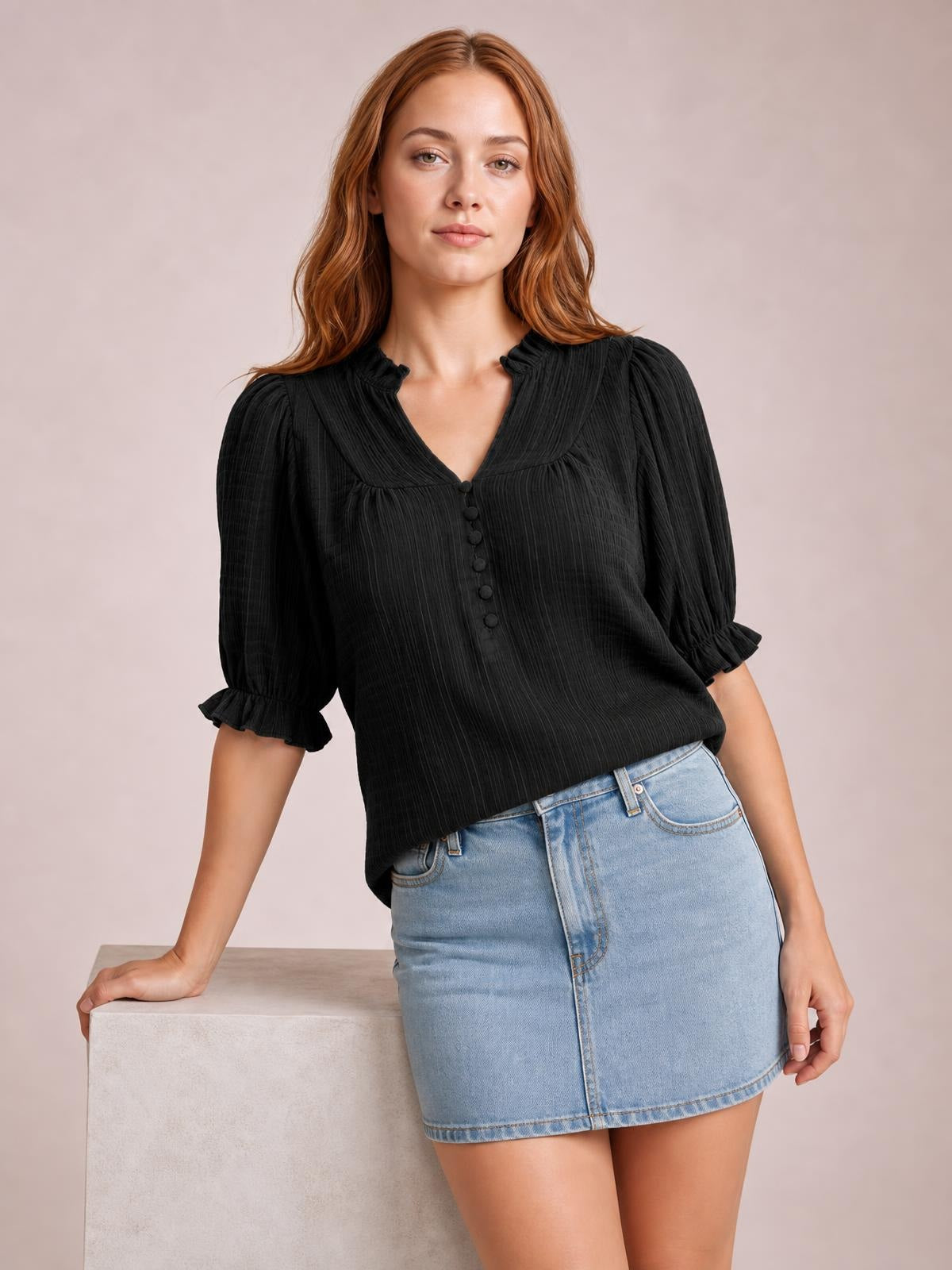 A model wears a black frill notched short sleeve blouse with ruched texture, button front, gathered cuffs, and a relaxed silhouette while leaning on a block.