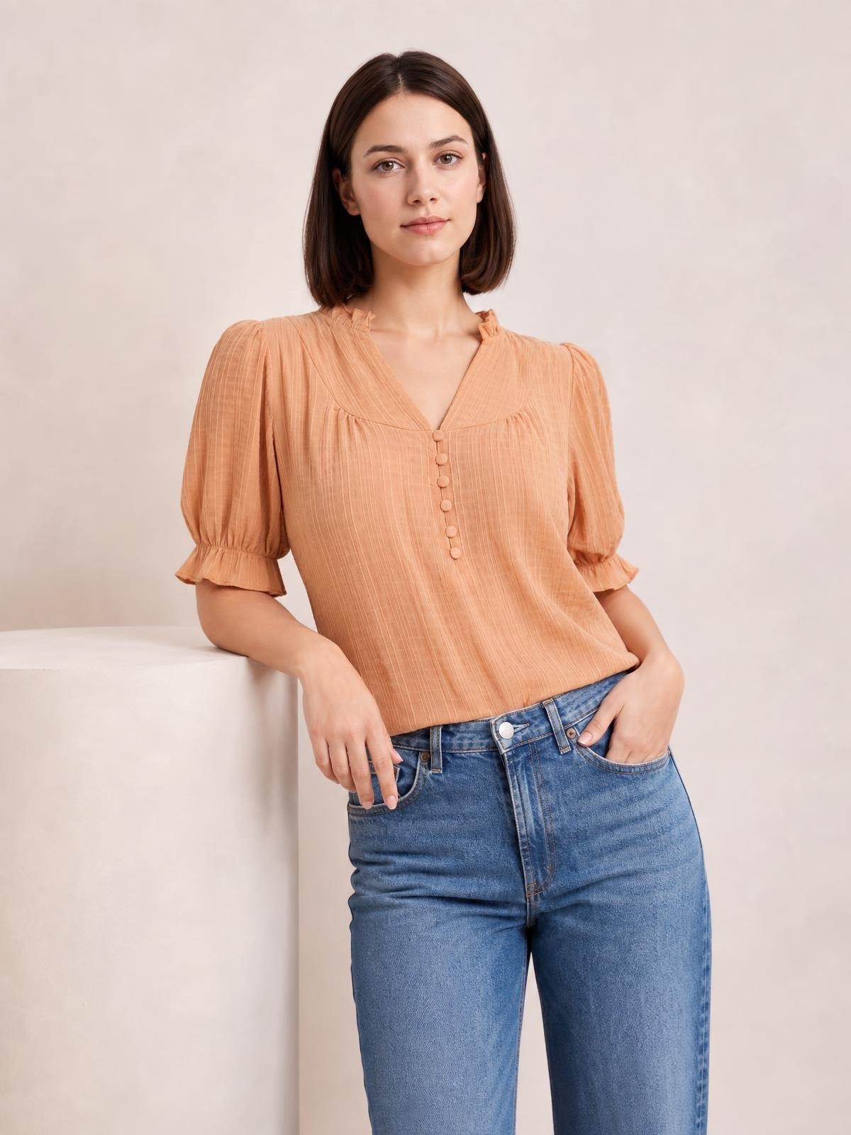 Model wears a peach frill notched short sleeve blouse with a V-neck, ruched yoke, button front, puffed sleeves, and ruffled cuffs in a relaxed silhouette.