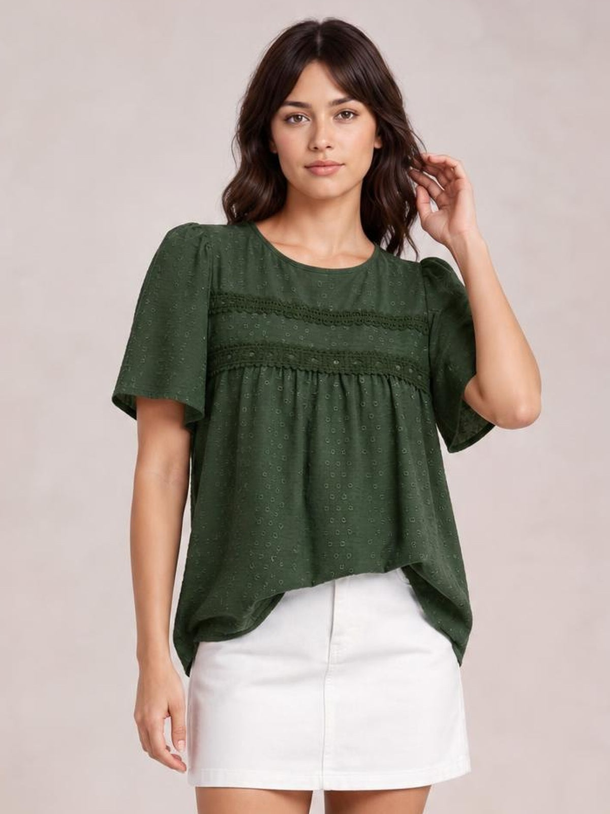 Model poses with one hand in her hair wearing a dark green lace detail round neck blouse with short sleeves, ruched trim, and a loose flowy silhouette.