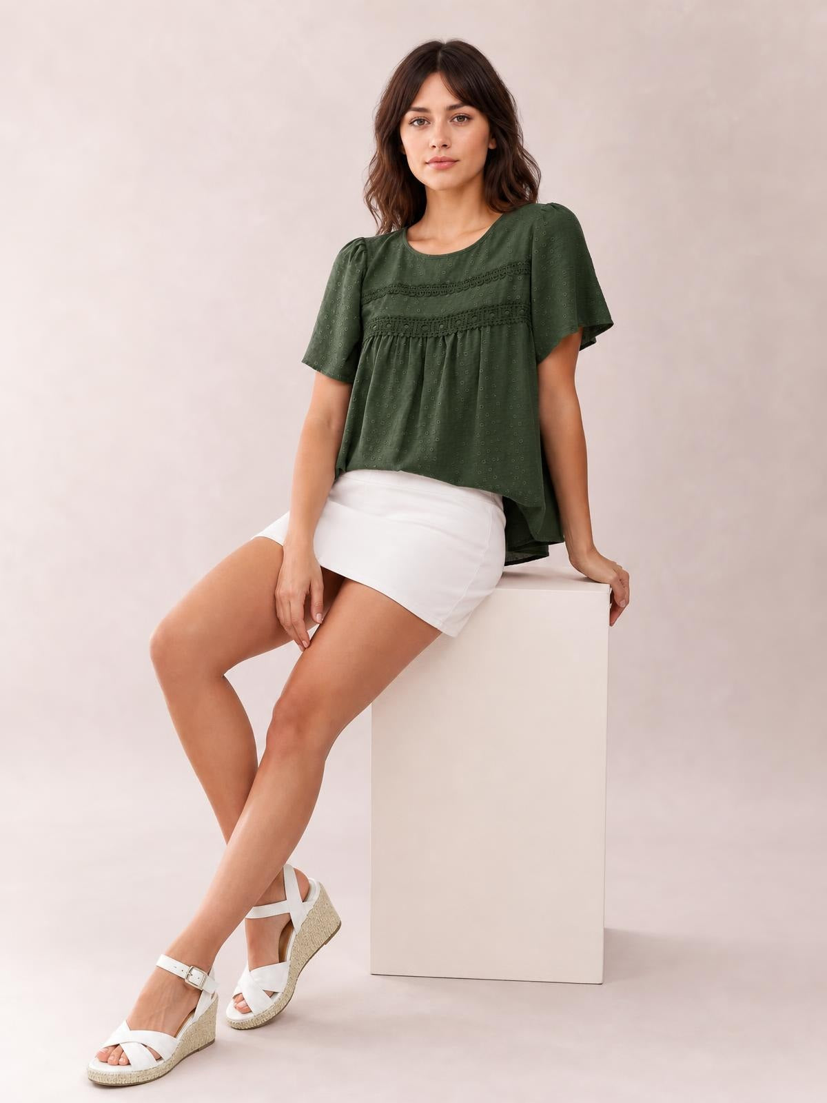 A seated model wears a dark green round-neck short-sleeve blouse with lace detail, ruched bands, a relaxed silhouette, and a gently flared hem.