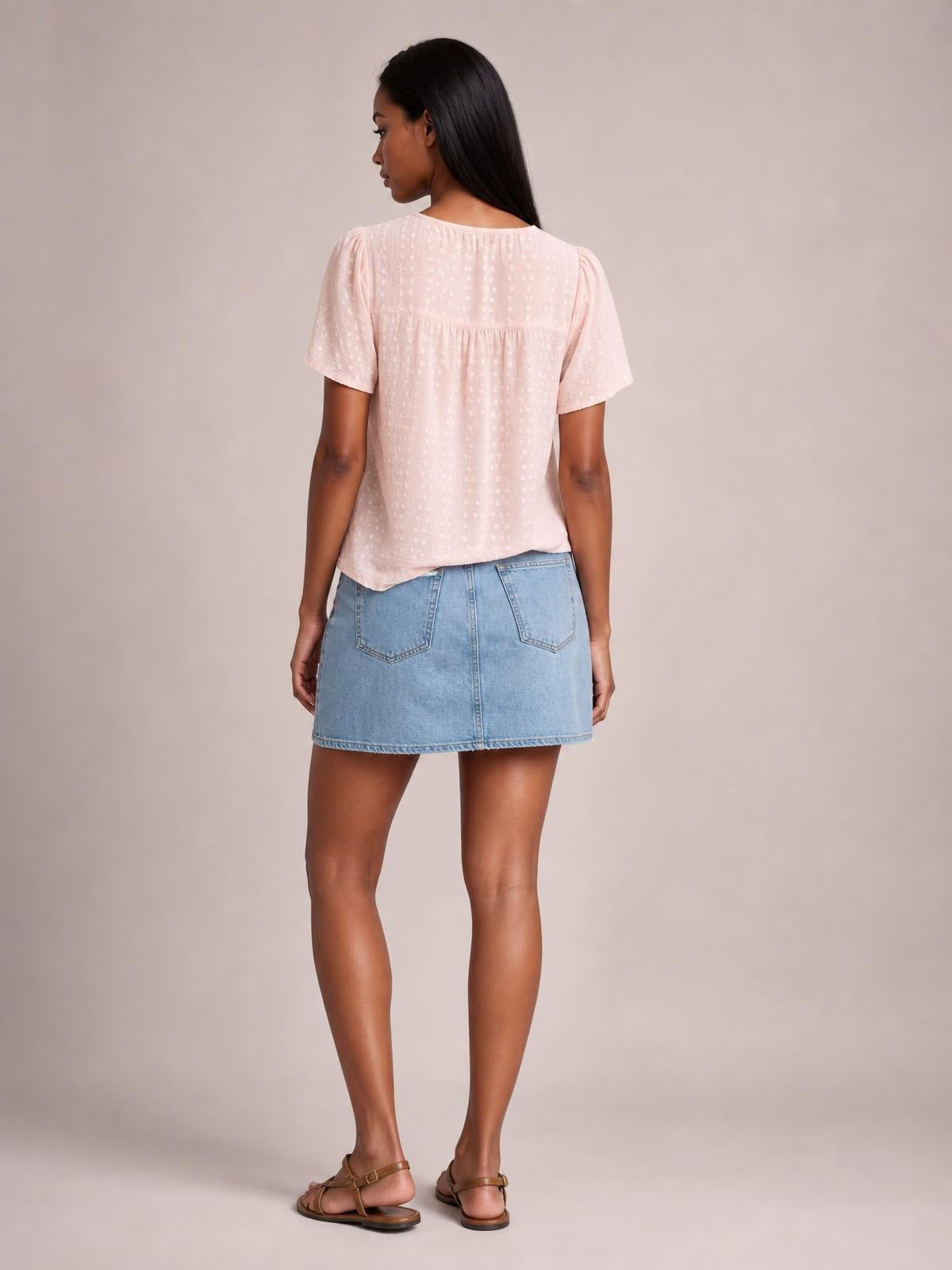 A model stands with her back to the camera in a pale pink lace-detail blouse with a round neck, short sleeves, and a softly ruched yoke.