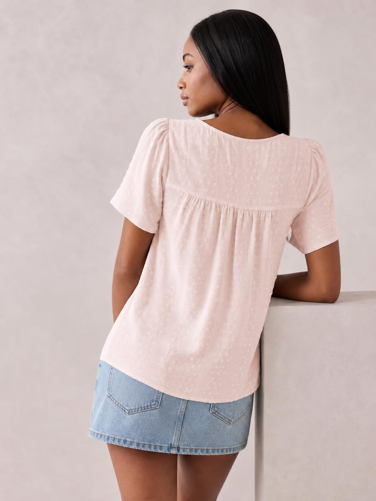 Shown from the back, a model wears a pale pink round-neck short-sleeve blouse with lace-textured fabric, ruched gathering below the yoke, and a relaxed silhouette.