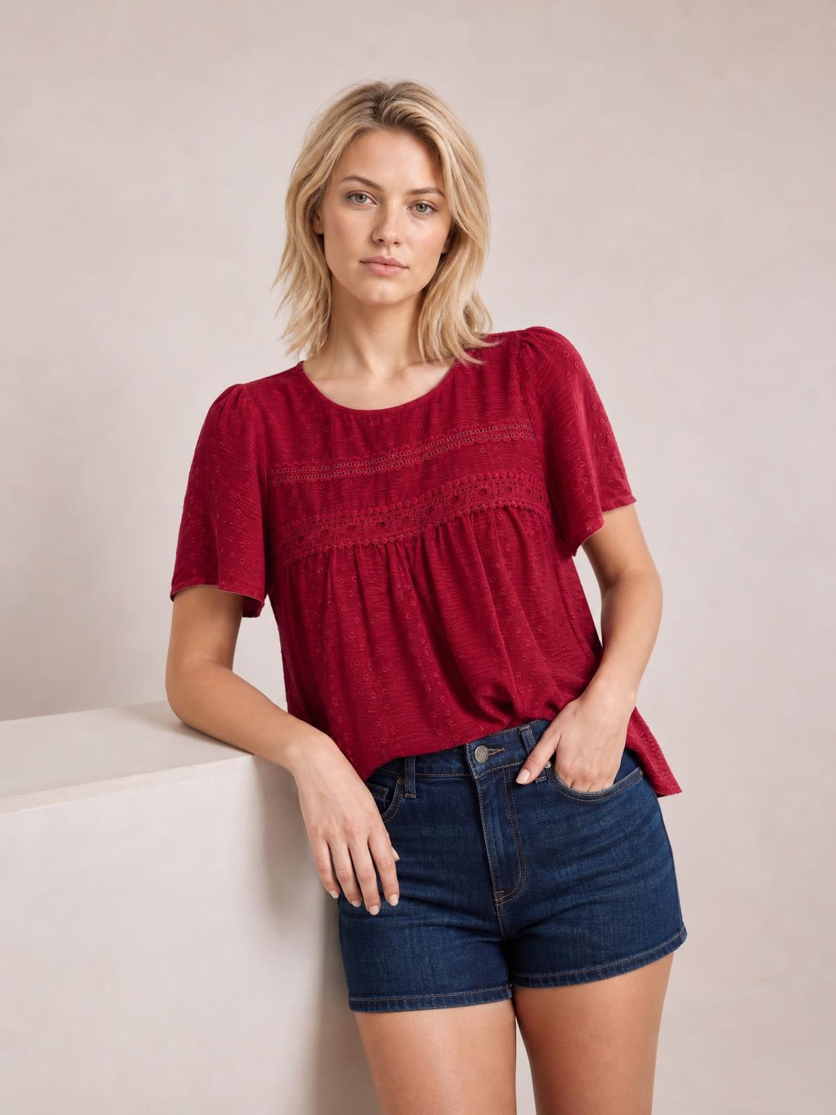 A model leans casually in a burgundy round-neck short-sleeve blouse with lace detail, ruched gathers, and a relaxed flowing silhouette.