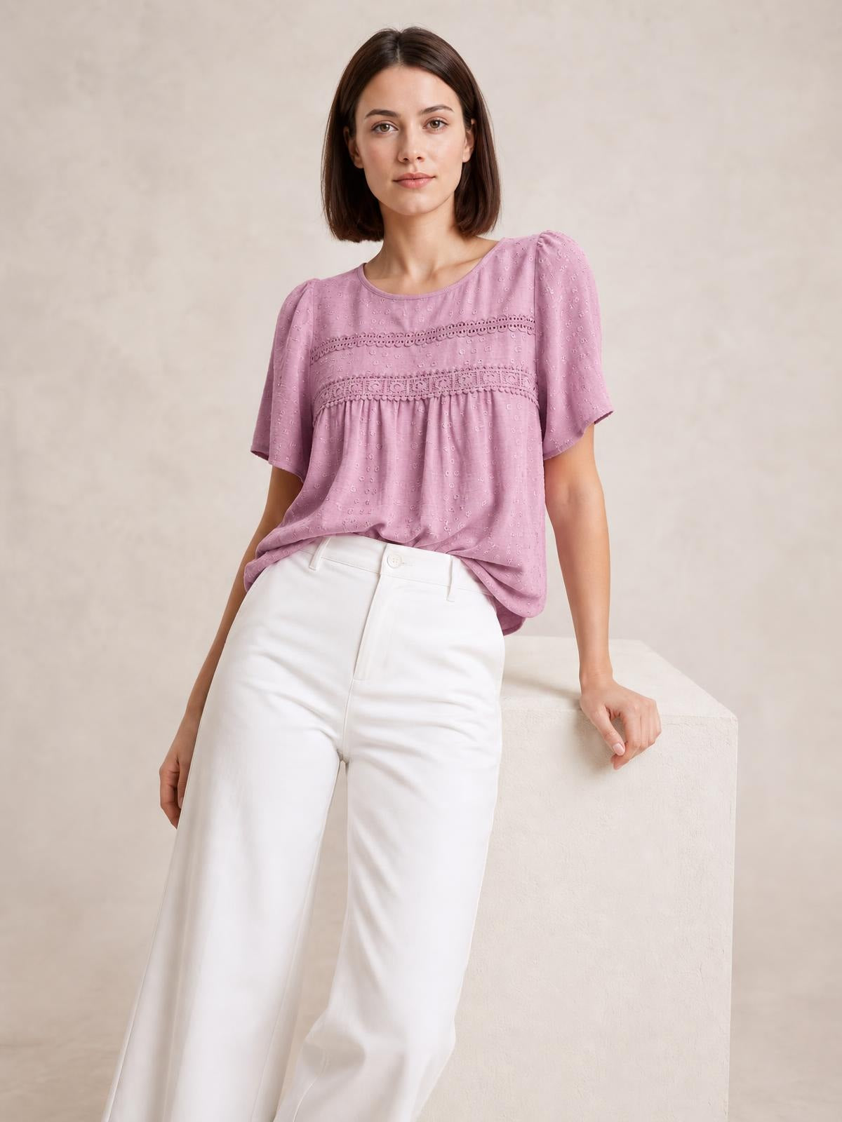 Model wearing a dusty pink lace detail round neck blouse with short sleeves, ruched waist, and a relaxed silhouette while leaning against a ledge.