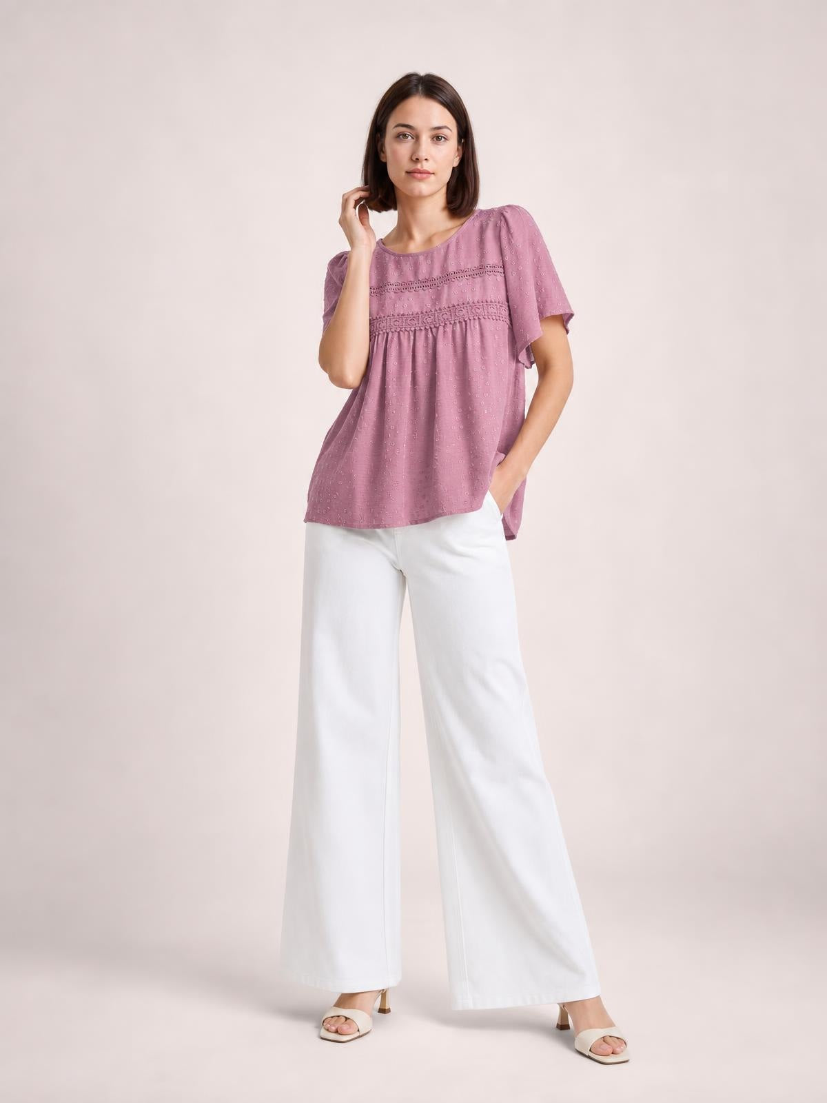 Model wearing a mauve round-neck short-sleeve blouse with lace trim, ruched details, a relaxed flowing silhouette, and a slightly flared hem.