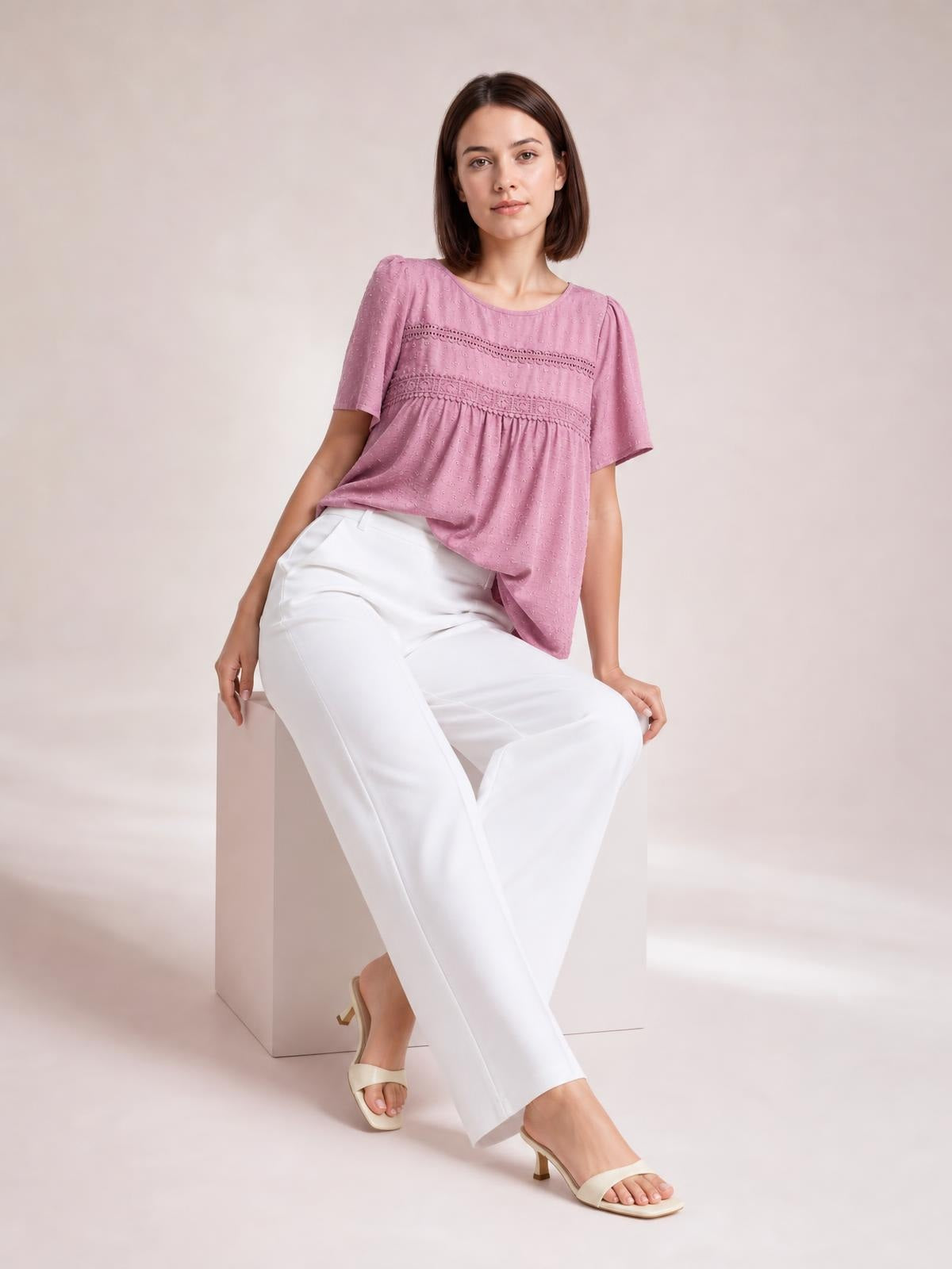 Model seated in a pink round-neck short-sleeve blouse with lace detail and ruched seams, featuring a loose, flowing silhouette.