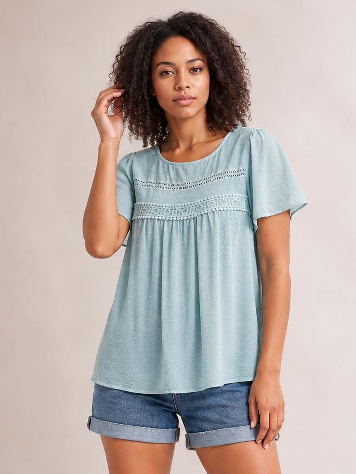 A model poses with one hand in her hair, wearing a pale blue round-neck short-sleeve blouse with lace trim, ruched gathers, and a loose silhouette.