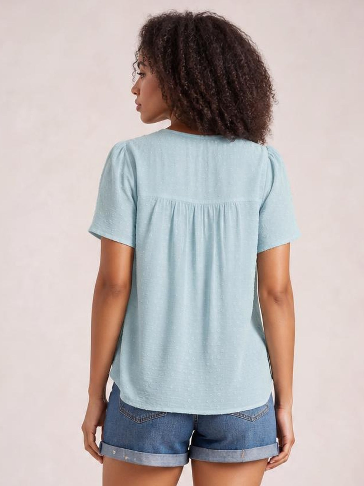 A model faces away wearing a light blue round-neck short-sleeve blouse with a relaxed silhouette, gathered ruched back yoke, and curved hem.