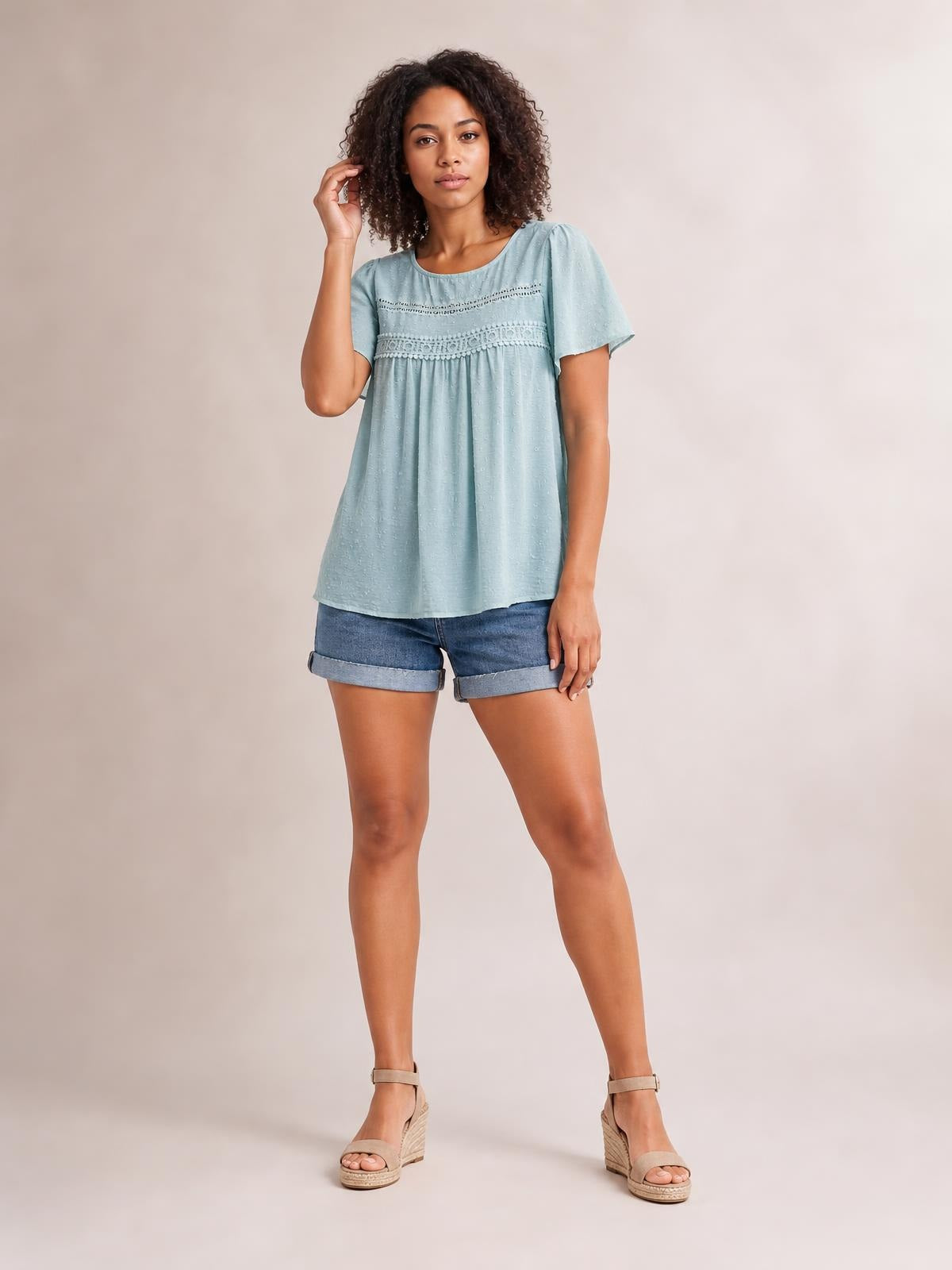Model poses with one hand in her hair wearing a light blue round-neck short-sleeve blouse with lace trim, ruched detailing, and a loose flowing silhouette.