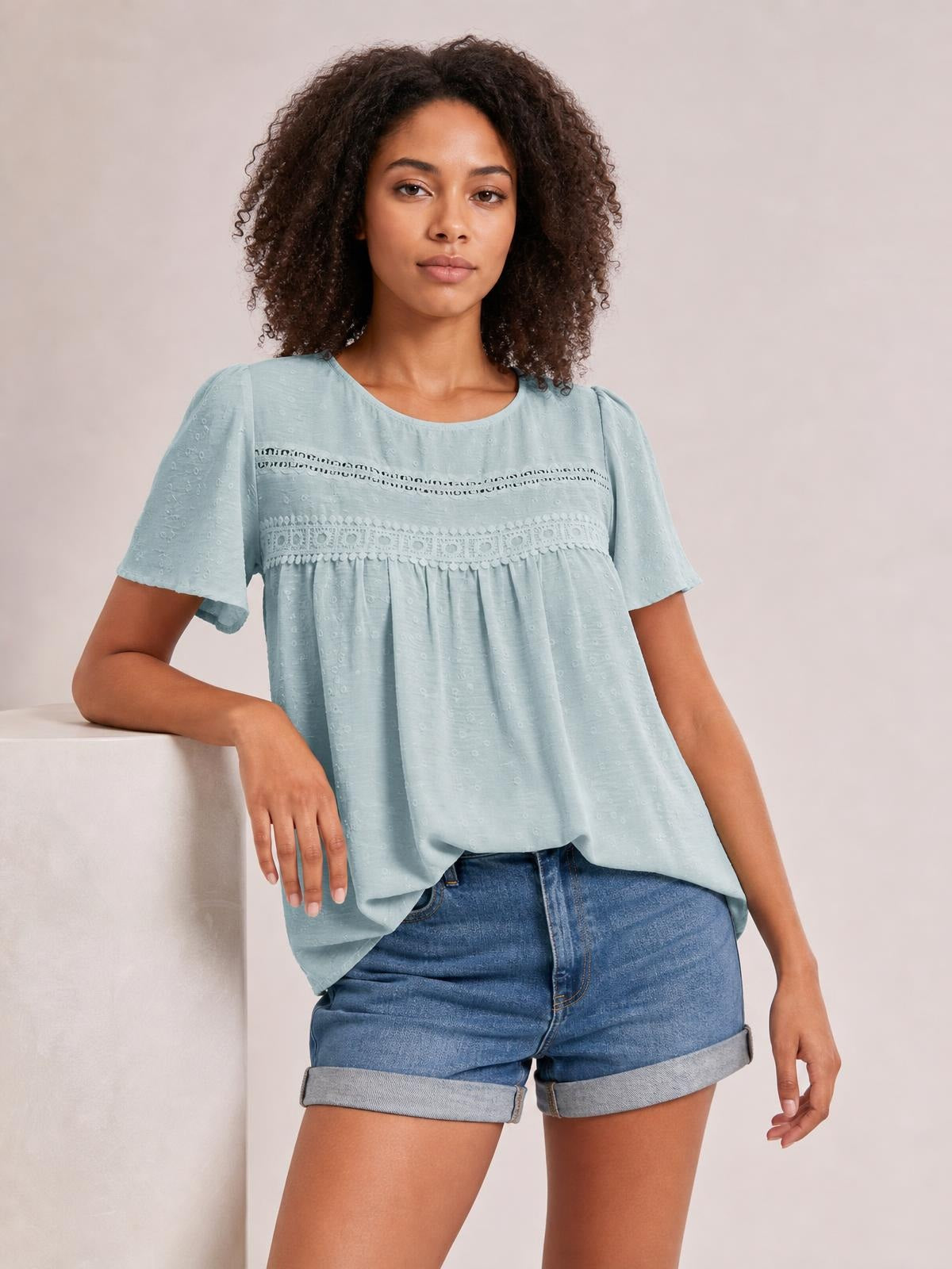 A model wears a pale blue round-neck short-sleeve blouse with lace detail, ruched gathering, and a relaxed, flowy silhouette while leaning on a ledge.
