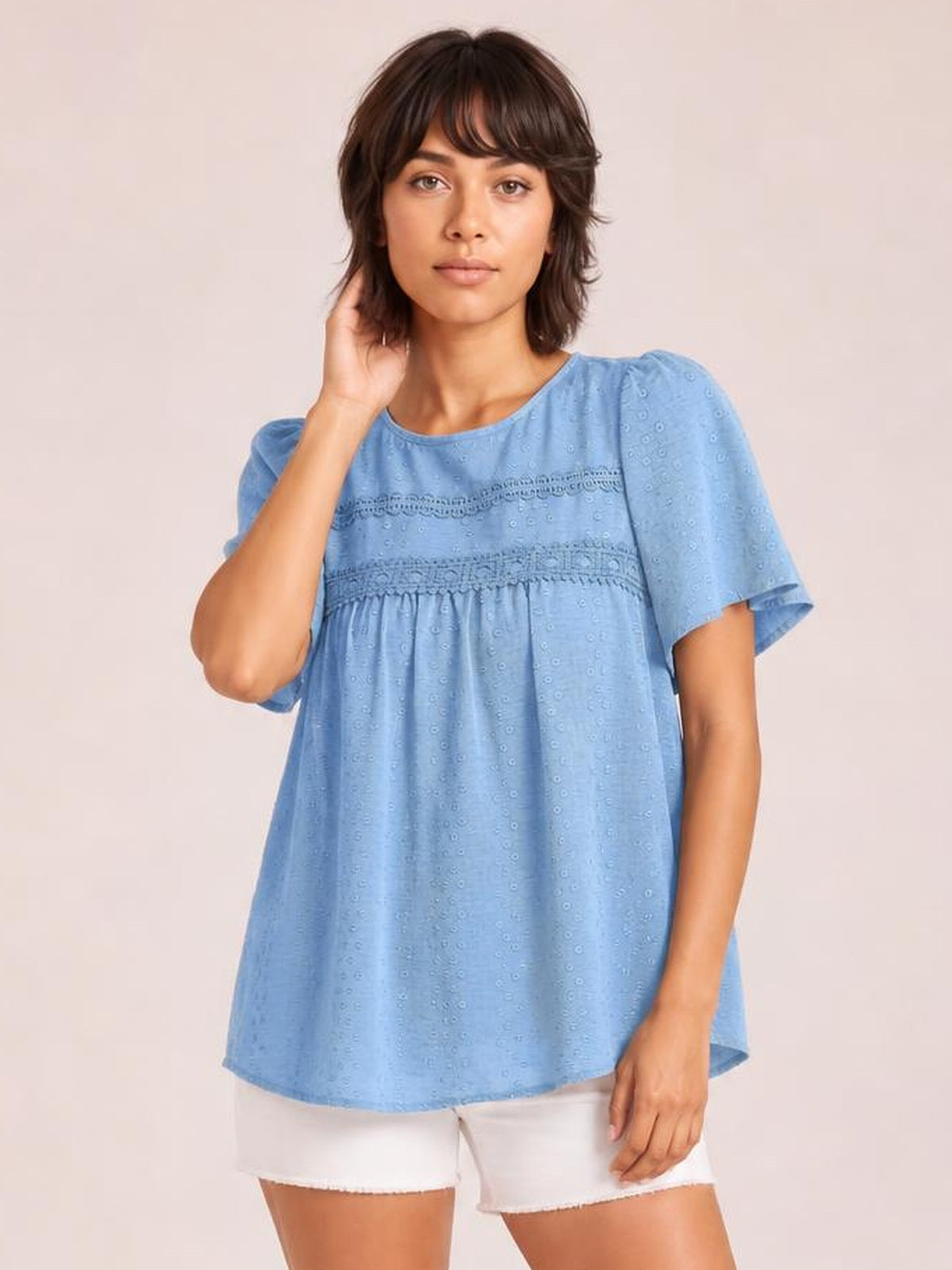 Model poses with one hand at her hair wearing a light blue round-neck blouse with short sleeves, lace bands, ruched detailing, and a relaxed babydoll silhouette.