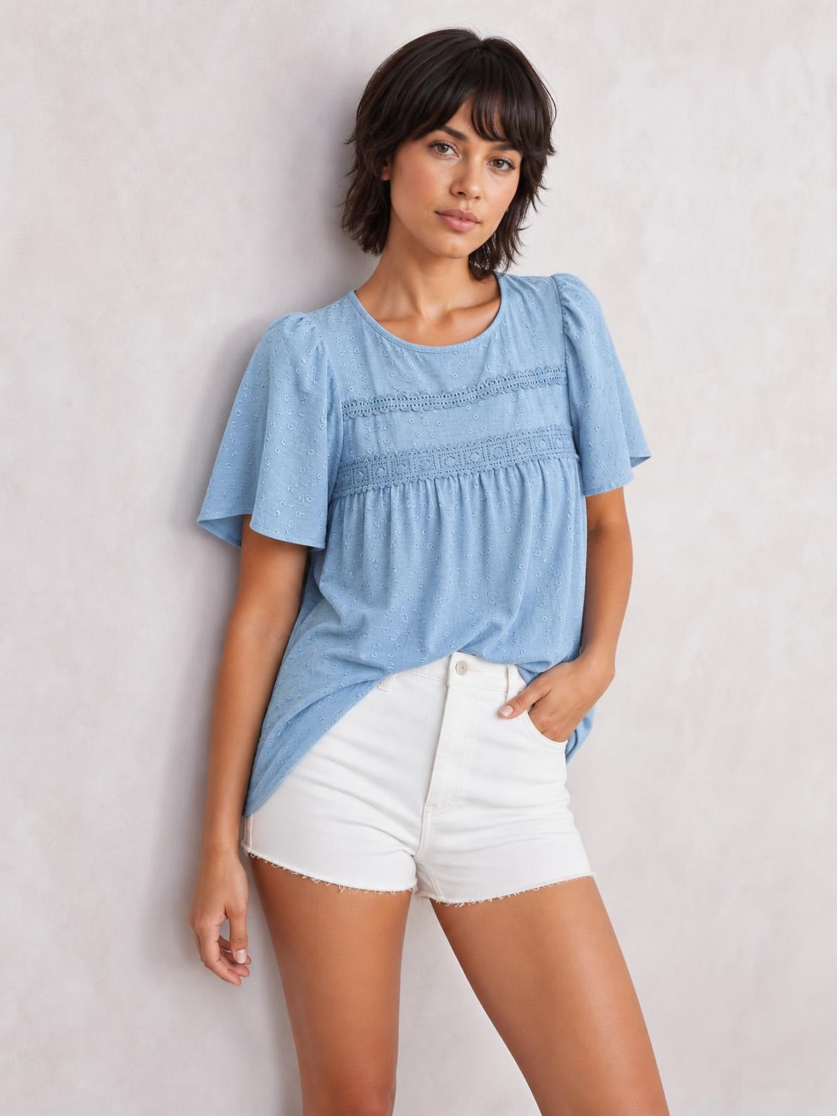 Model poses in a light blue lace detail blouse with a round neck, short flutter sleeves, ruched horizontal trim, and a relaxed silhouette.