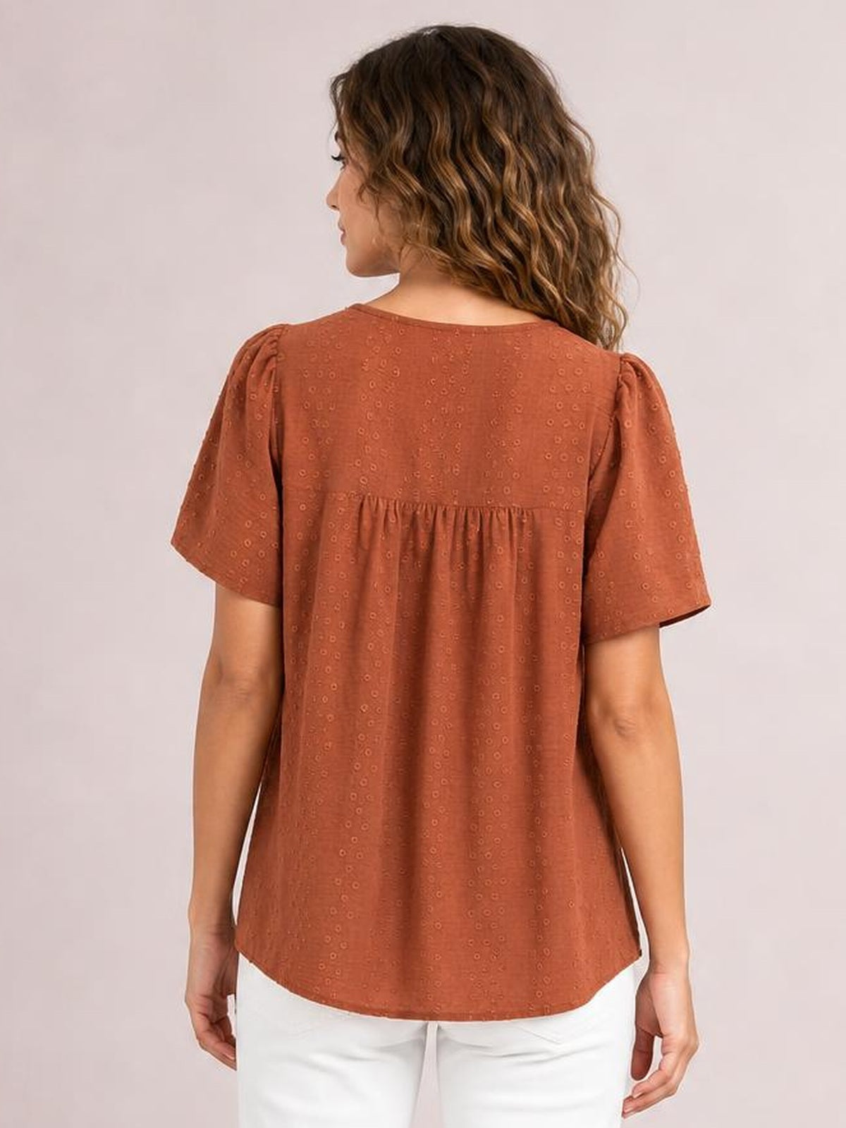 A model shown from behind wears a rust-colored round-neck blouse with short sleeves, lace detail, ruched back yoke, and a relaxed silhouette.