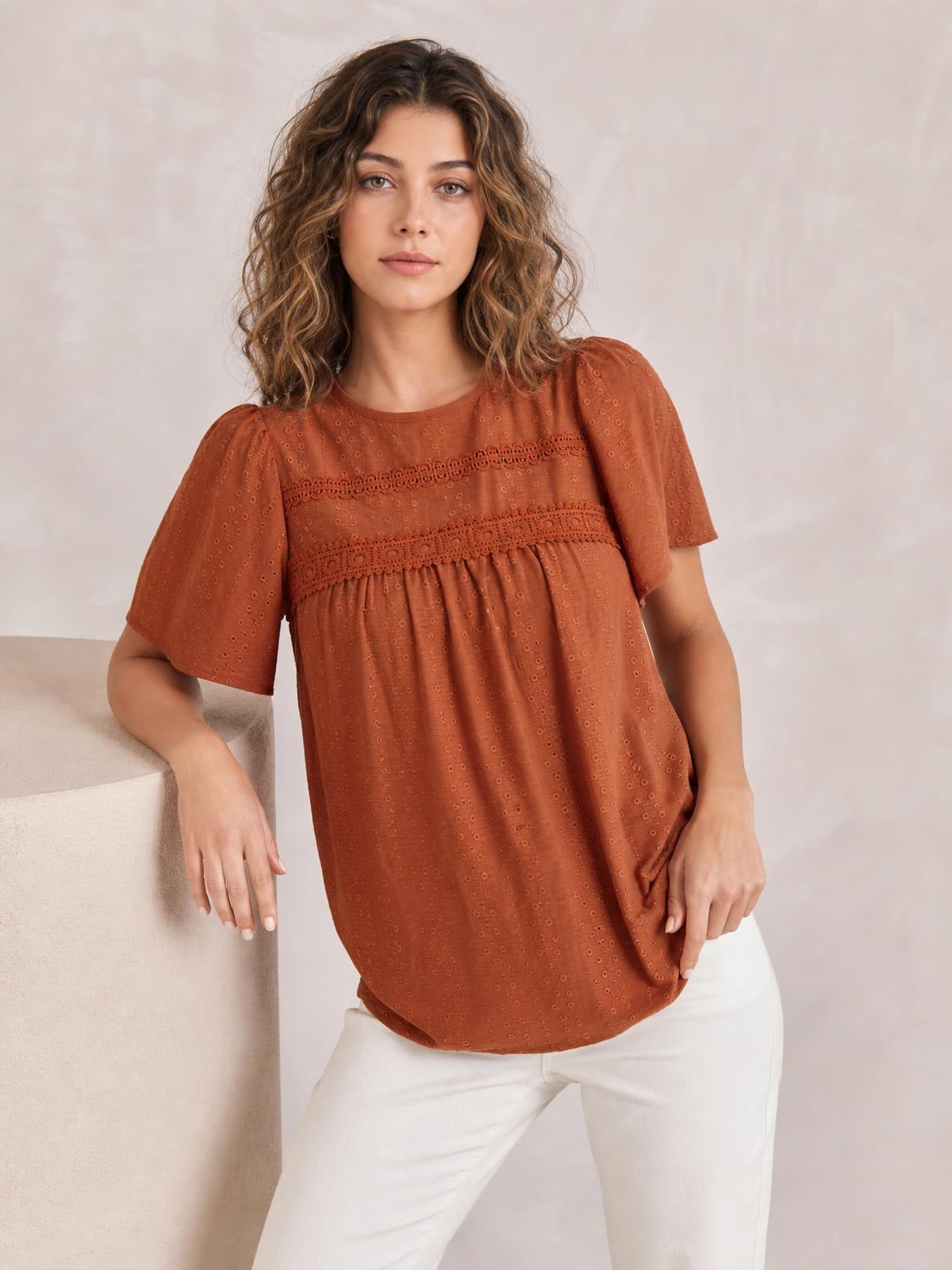 A model wears a rust-brown round-neck short-sleeve blouse with lace trim, ruched detailing, and a relaxed flowing silhouette while standing.