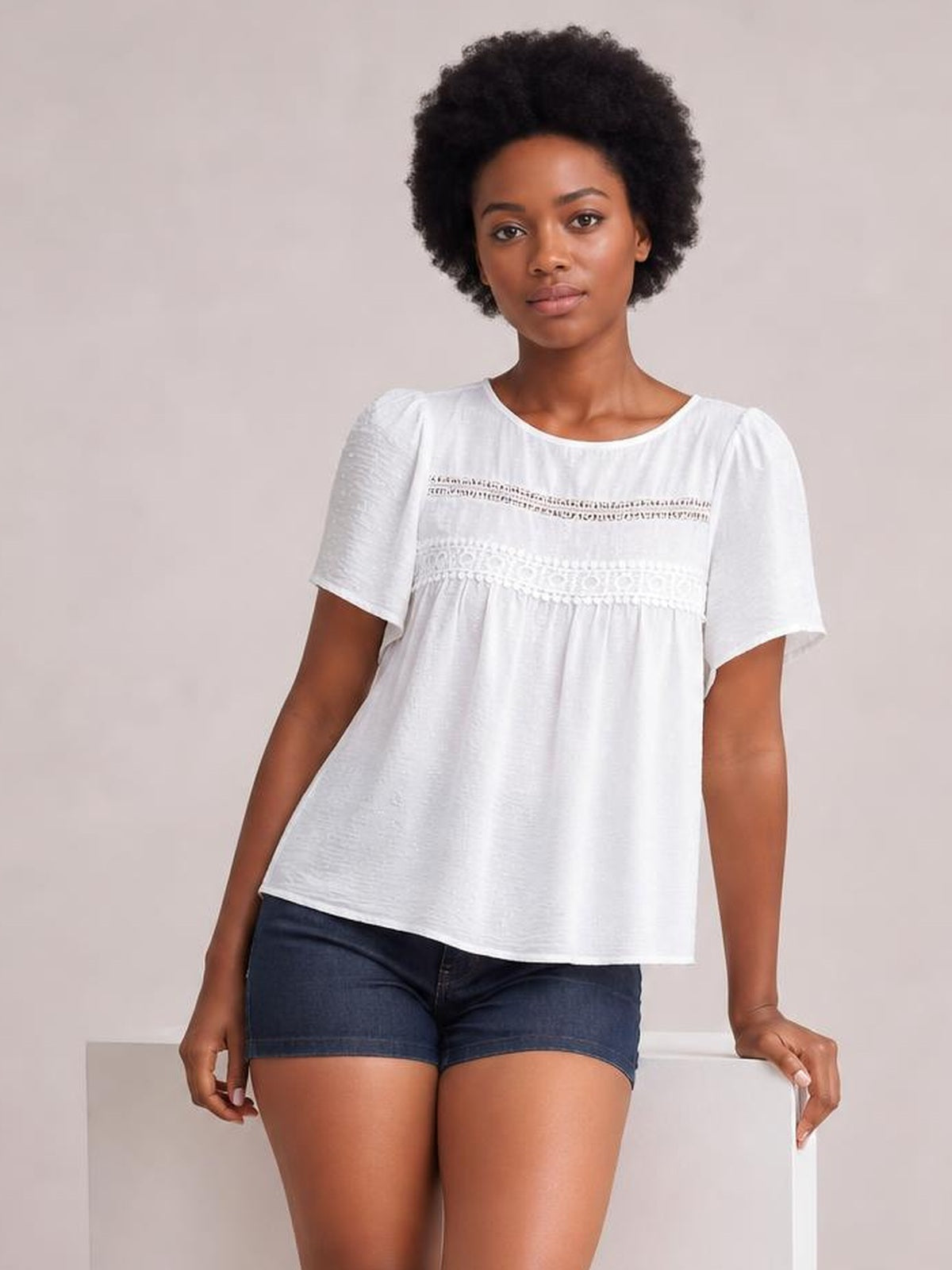 A model wears a white round-neck short-sleeve blouse with lace trim, ruched detailing, gathered shoulders, and a relaxed flowing silhouette.