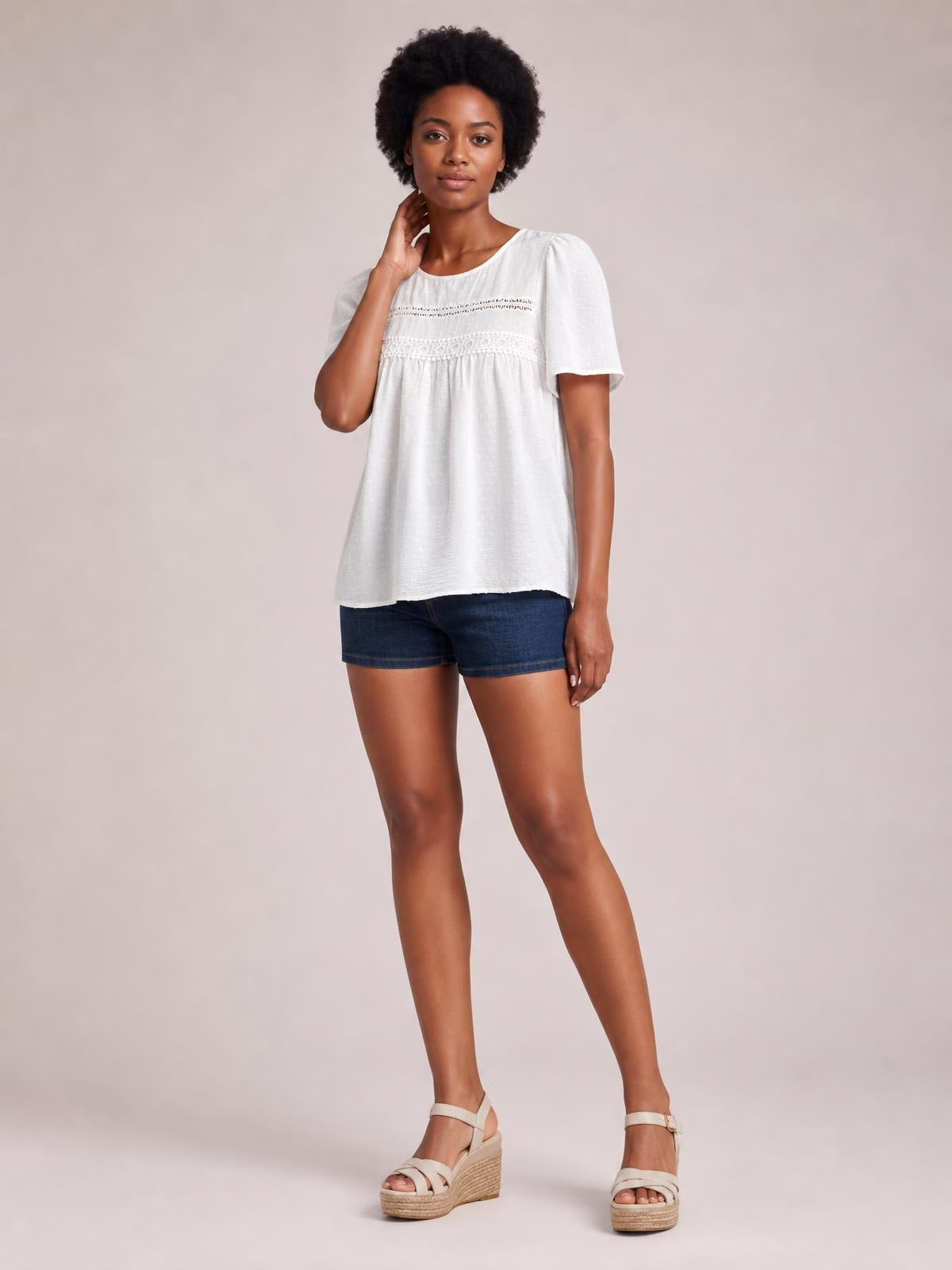 A model stands in a white round-neck short-sleeve blouse with lace trim and ruched detailing, featuring a relaxed, softly flared silhouette.