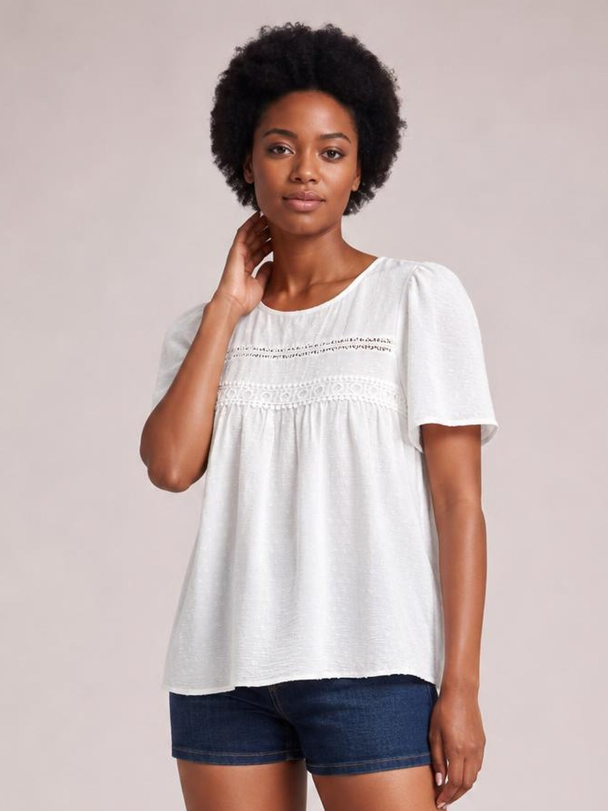Model wearing a white round-neck short-sleeve blouse with lace and ruched chest details, a relaxed flared silhouette, and a curved hem.