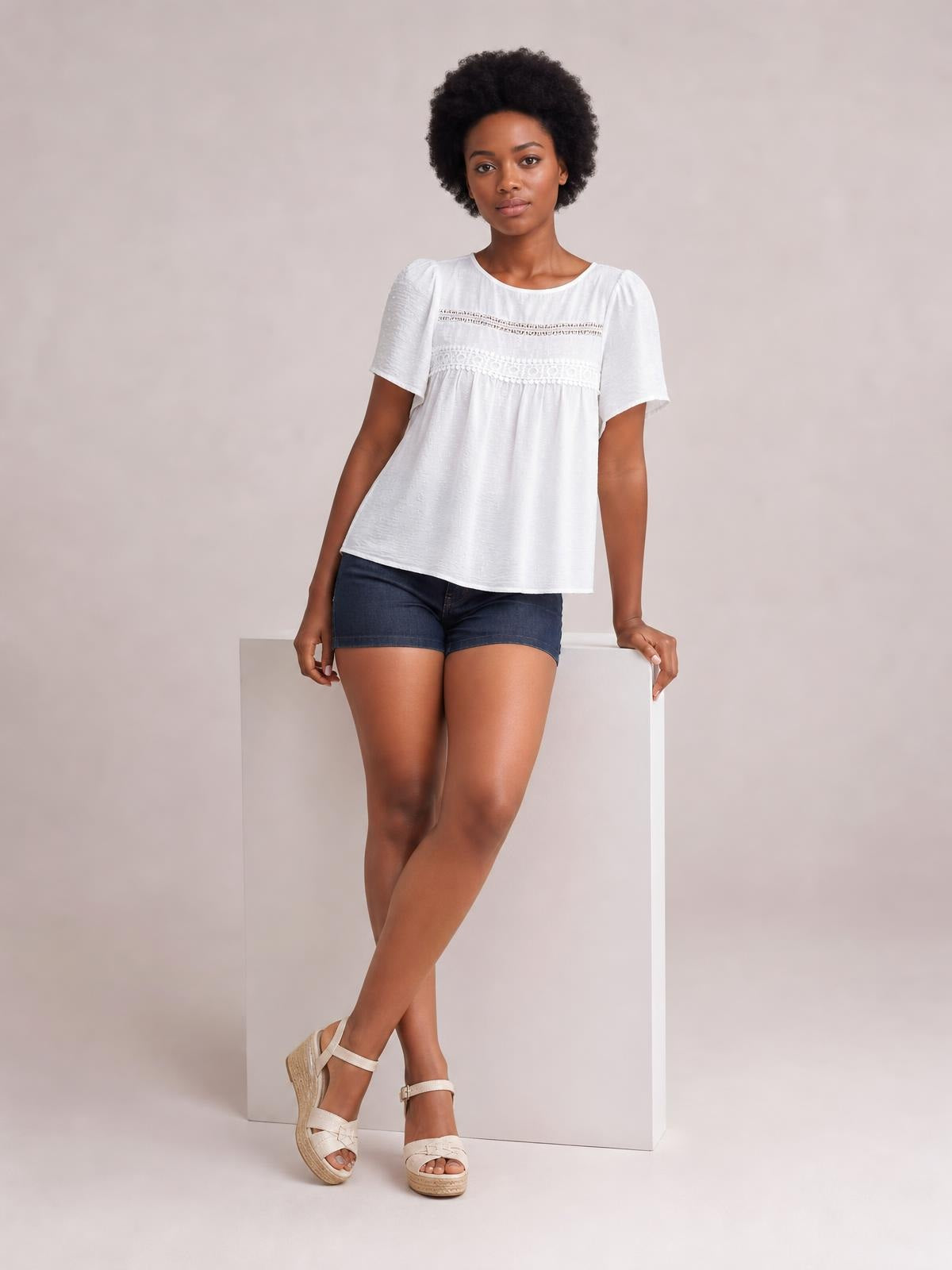 Model wears a white round-neck short-sleeve blouse with lace detail, ruched seams, and a relaxed, flowy silhouette while leaning against a pedestal.