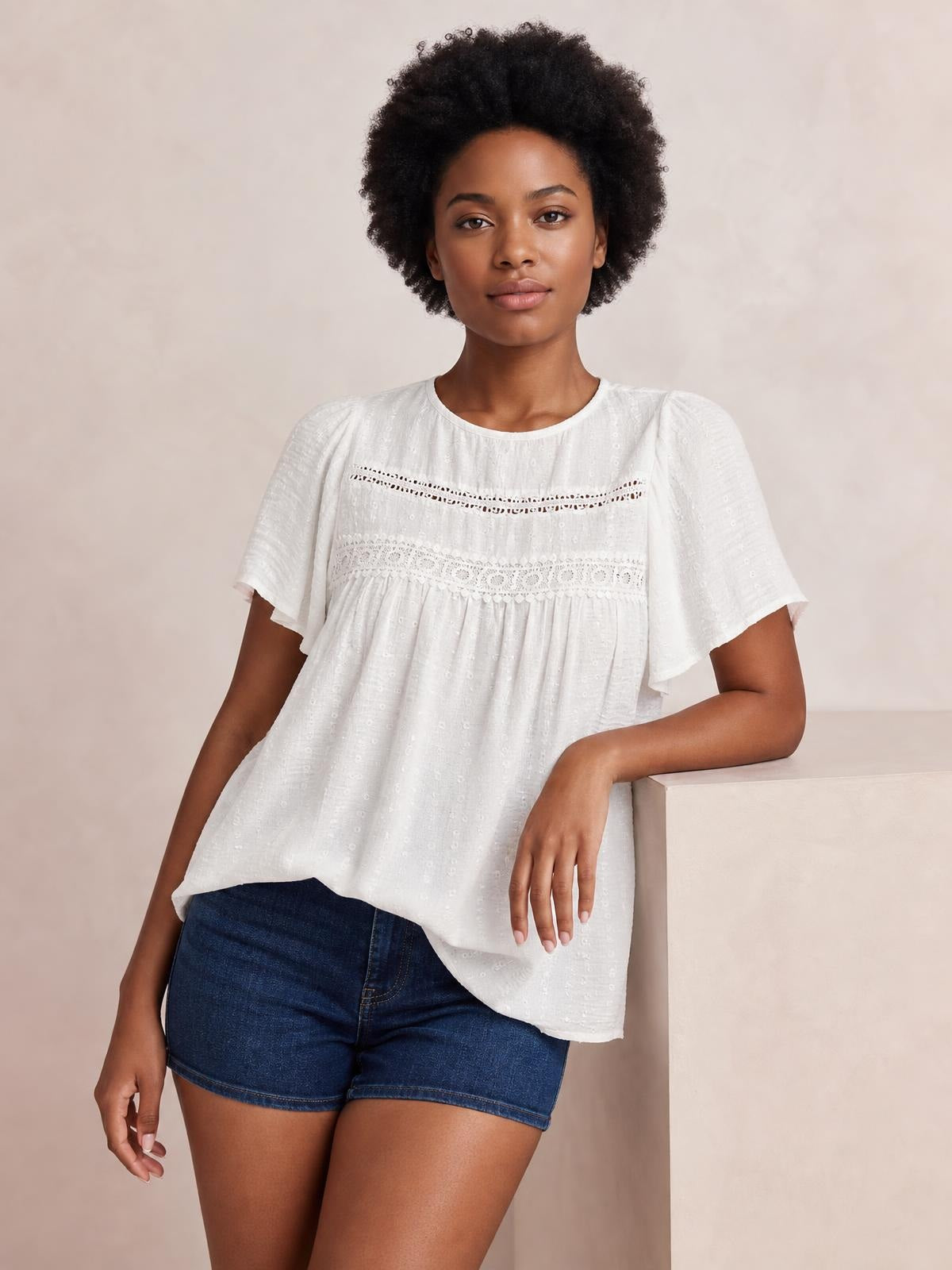 Model poses in a white round-neck short-sleeve blouse with lace detail, ruched seams, fluttered sleeves, and a loose flowing silhouette.