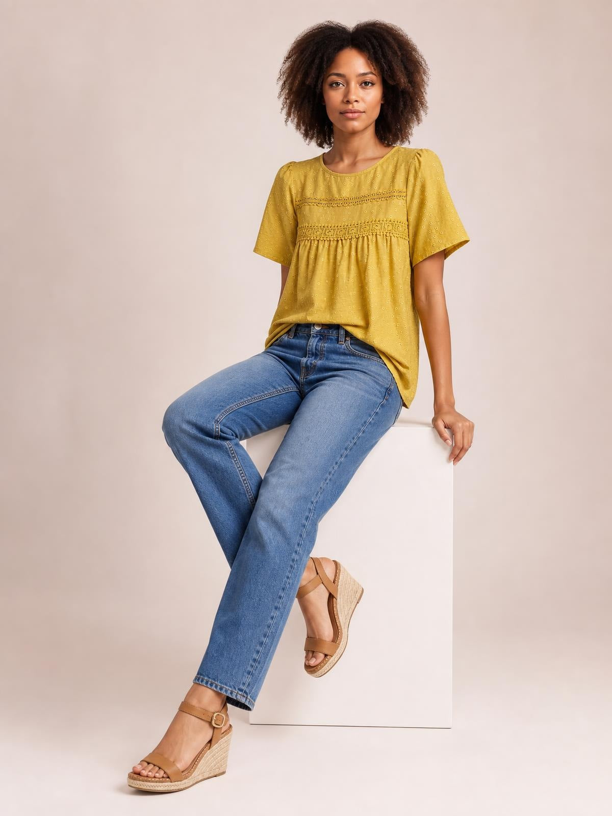A seated model wears a mustard-yellow round-neck short-sleeve blouse with lace trim, ruched gathering, and a relaxed silhouette.