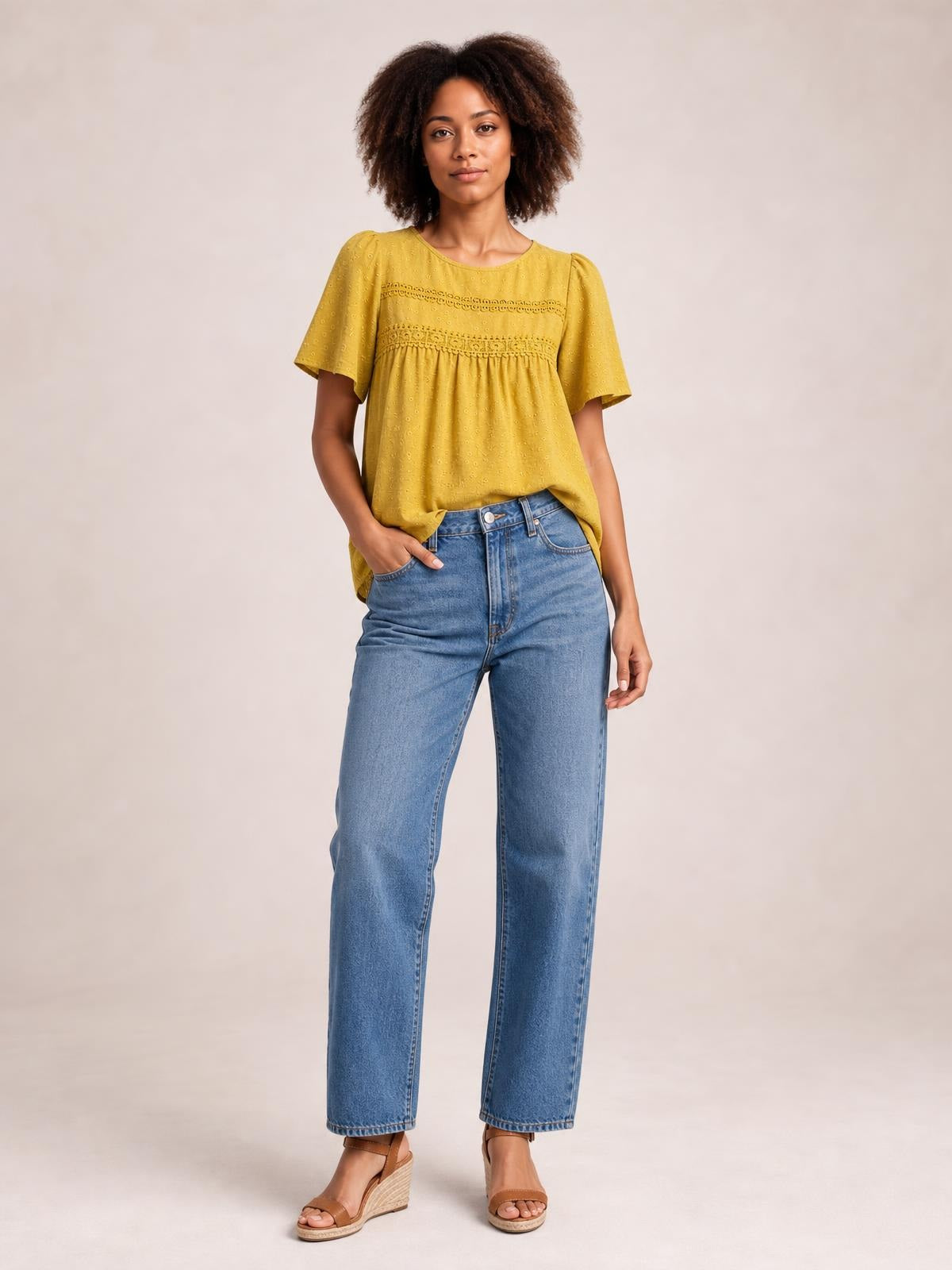 A model stands with one hand in her pocket wearing a mustard yellow round-neck short-sleeve blouse with lace trim, ruched detailing, and a relaxed silhouette.
