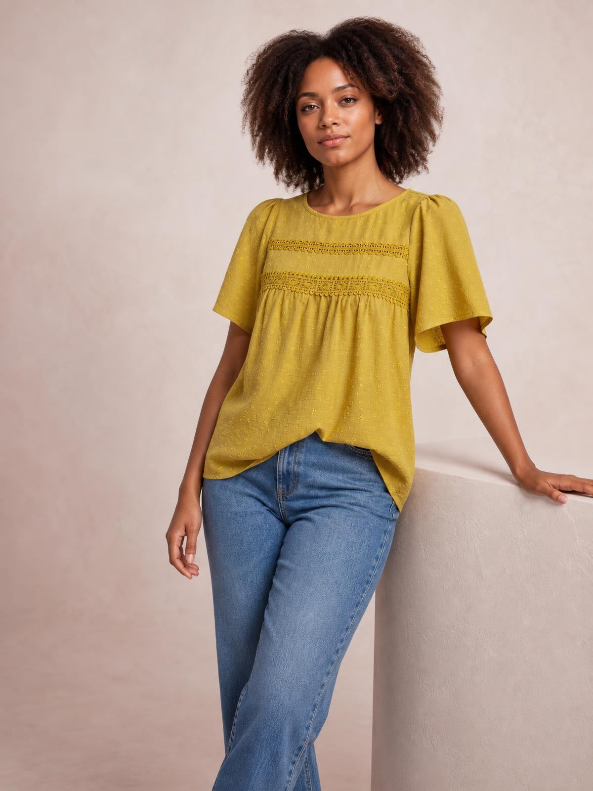 A model poses in a mustard yellow round-neck short-sleeve blouse with horizontal lace and ruched details, a relaxed flowing silhouette, and curved hem.