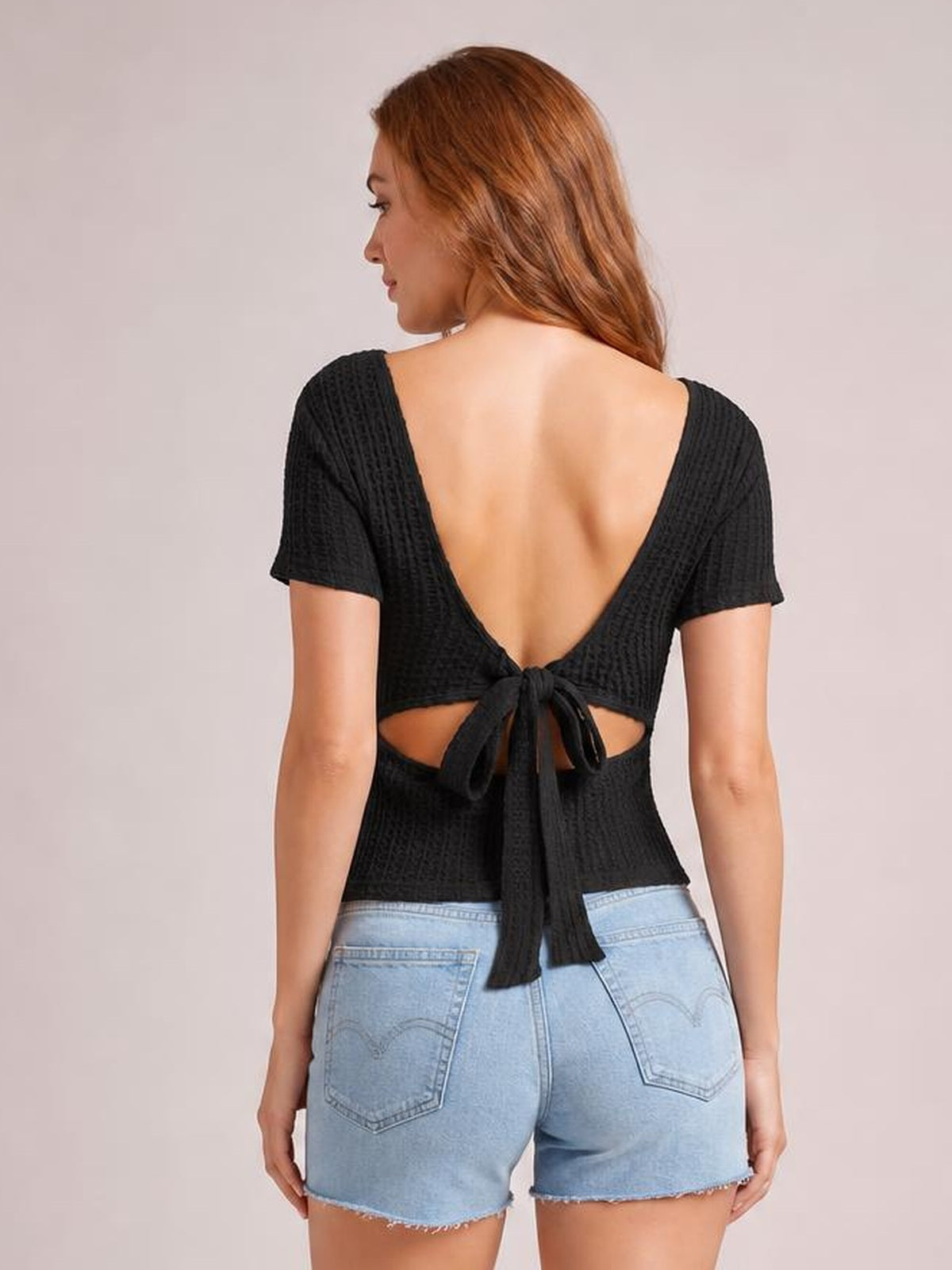 Model shown from behind wearing a black textured short-sleeve T-shirt with an open back, waist cutout, and long tied detail creating a fitted cropped silhouette.