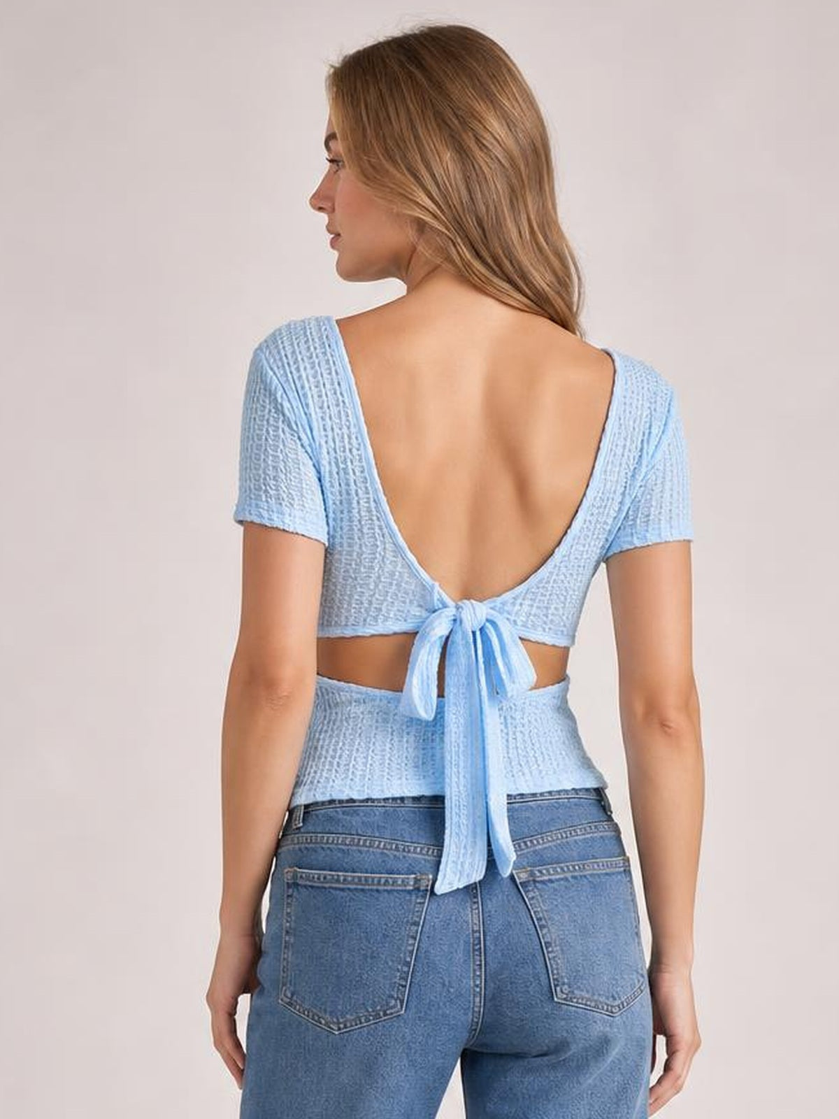 Model shown from the back wearing a light blue lace-detail short-sleeve T-shirt with a low square neckline, open-back silhouette, and tied waist detail.