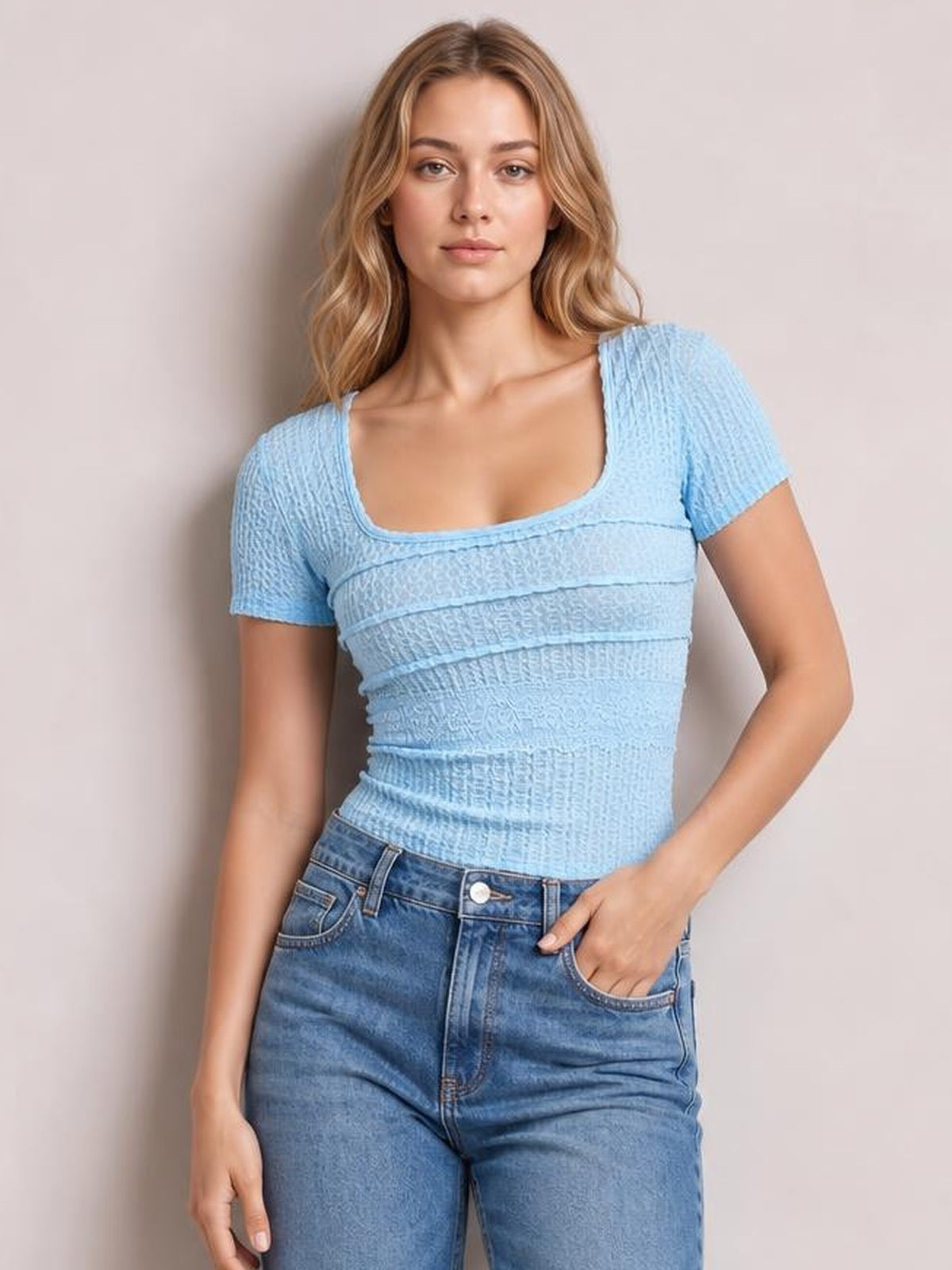 A model wears a fitted light blue lace-detail square-neck short-sleeve T-shirt with textured construction, posing with one hand in her jeans pocket.