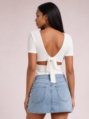 A model wears a white lace-detail short-sleeve T-shirt with a textured finish, open back, cropped silhouette, and tied detail at the waist.
